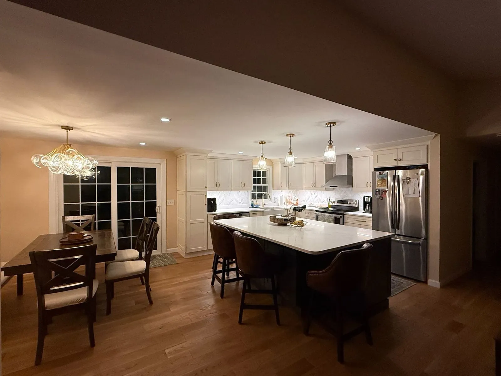 A remodeled kitchen at night with three glass pendants over a white island, glowing under-cabinet lights, recessed cans and a bubble-glass chandelier over the dining table.