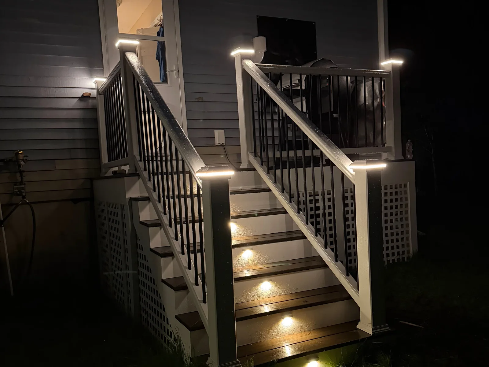Deck stairs at night lit by warm LED caps on each railing post and small lights on every stair riser, beside a lit back door.