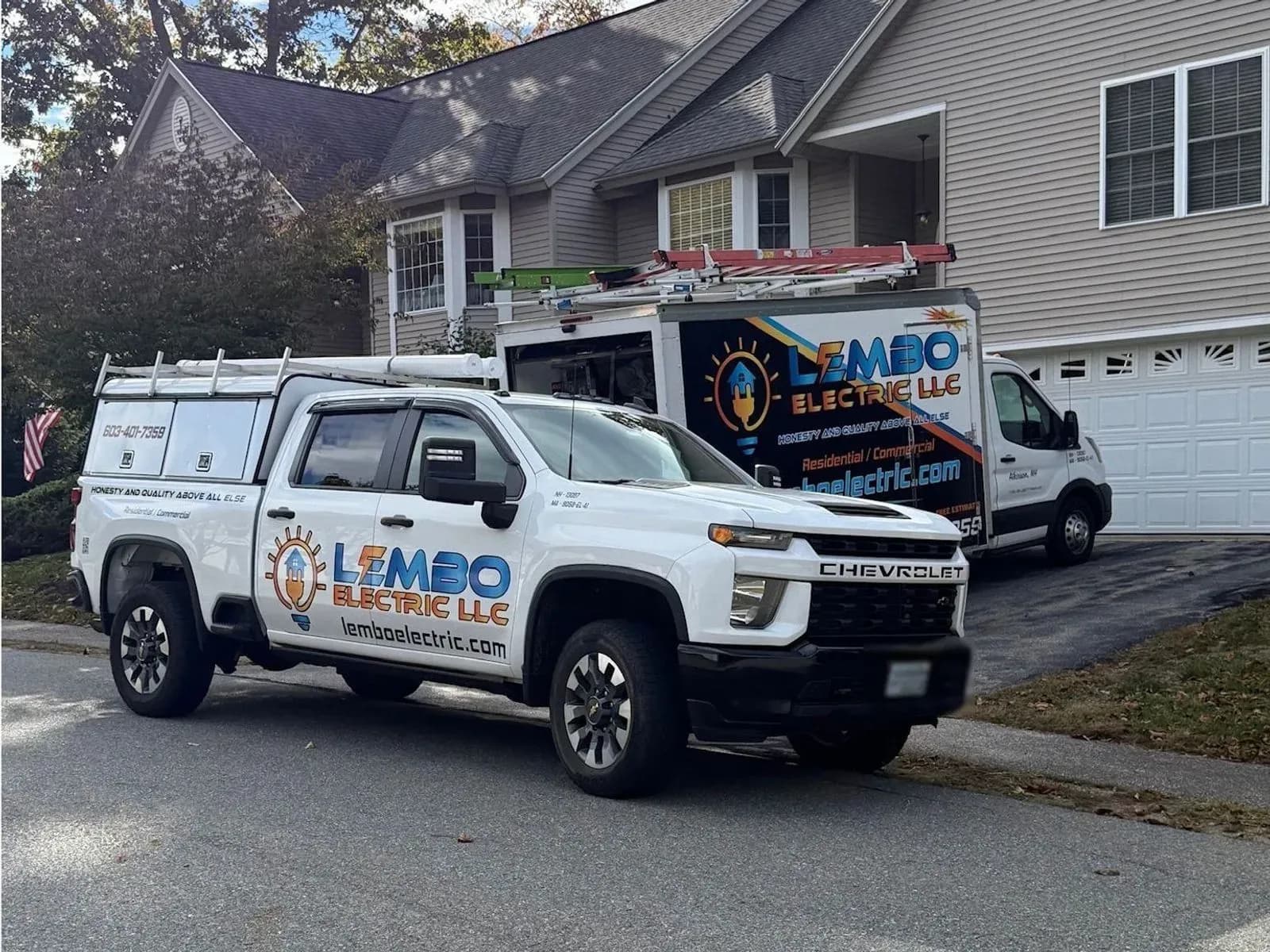 A white Lembo Electric pickup with a ladder rack parked at the curb and the lettered Lembo Electric box van in the driveway of a gray two-story house on a fall day.