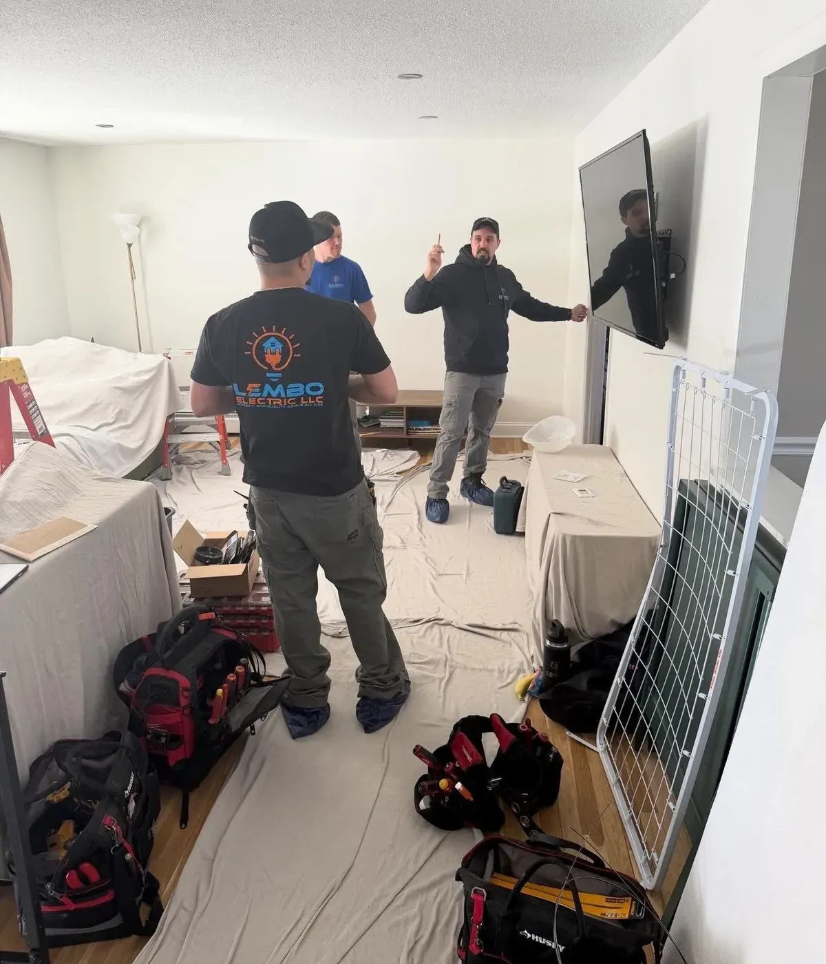 Three electricians, one in a Lembo Electric shirt, talk through a job in a living room with drop cloths over the floor and furniture, tool bags on the floor and a TV on the wall.