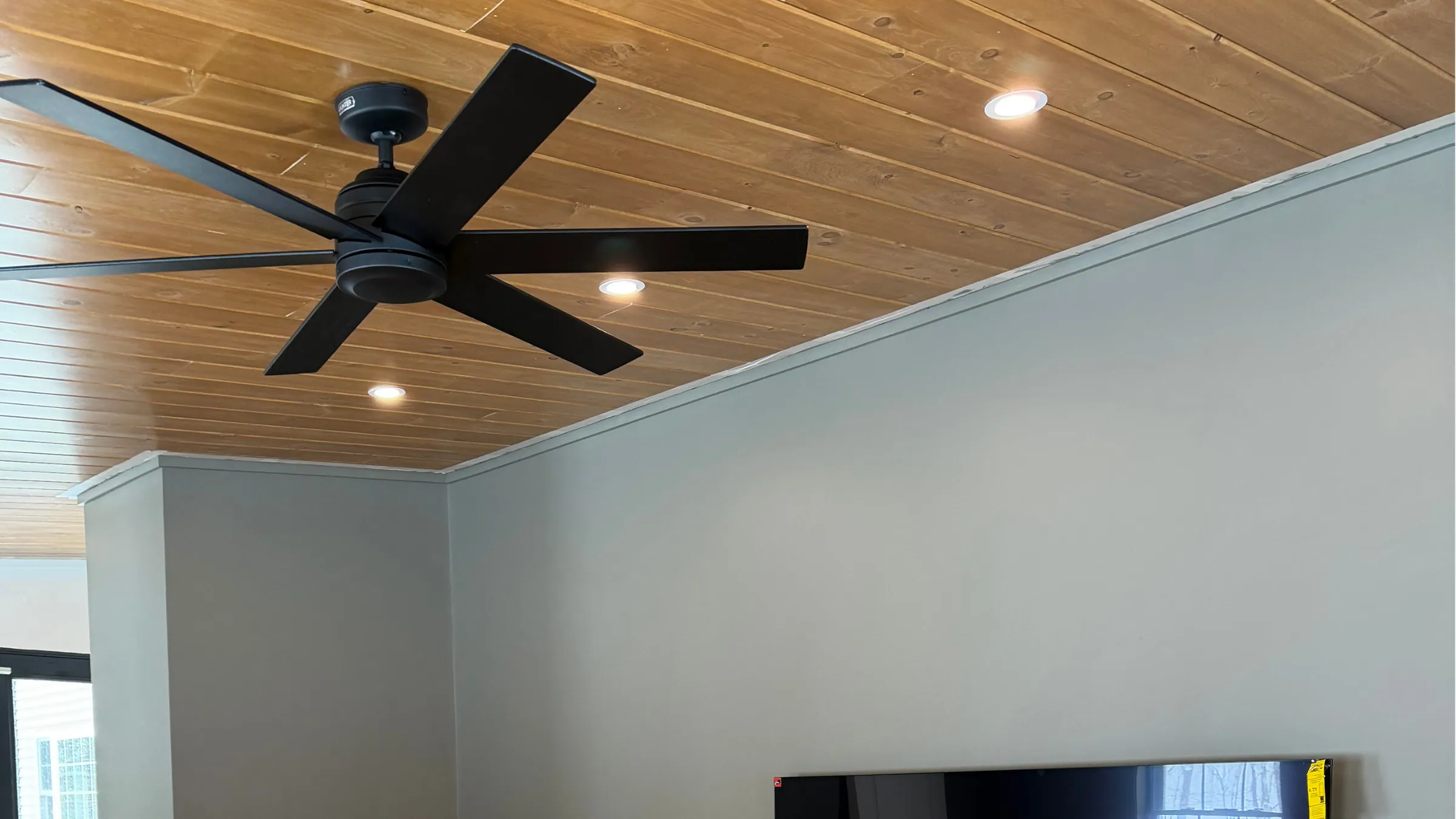 A black ceiling fan and recessed lights set in a knotty pine plank ceiling above gray walls, with a wall-mounted TV along the bottom edge.