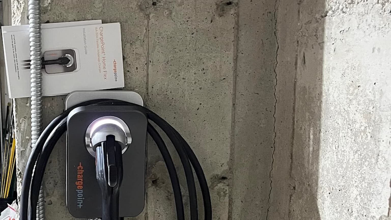 A ChargePoint Home Flex EV charger with its coiled black cable hung on a board-formed concrete wall, its installation guide tucked behind a run of flexible metal conduit.