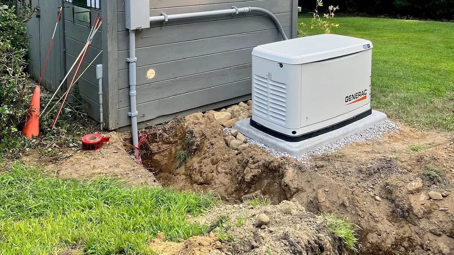 A new Generac standby generator on a gravel-edged pad beside a gray shed, a freshly dug trench still open in the soil in front of it.