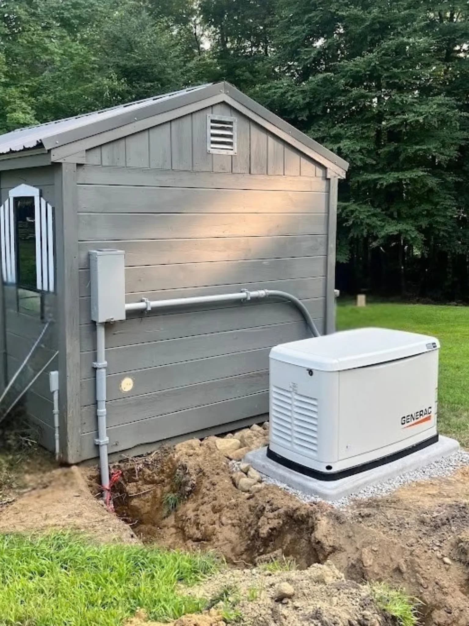 A new Generac standby generator on a gravel-edged pad beside a gray shed, conduit running from a disconnect on the shed wall, a freshly dug trench still open in the soil in front of it.