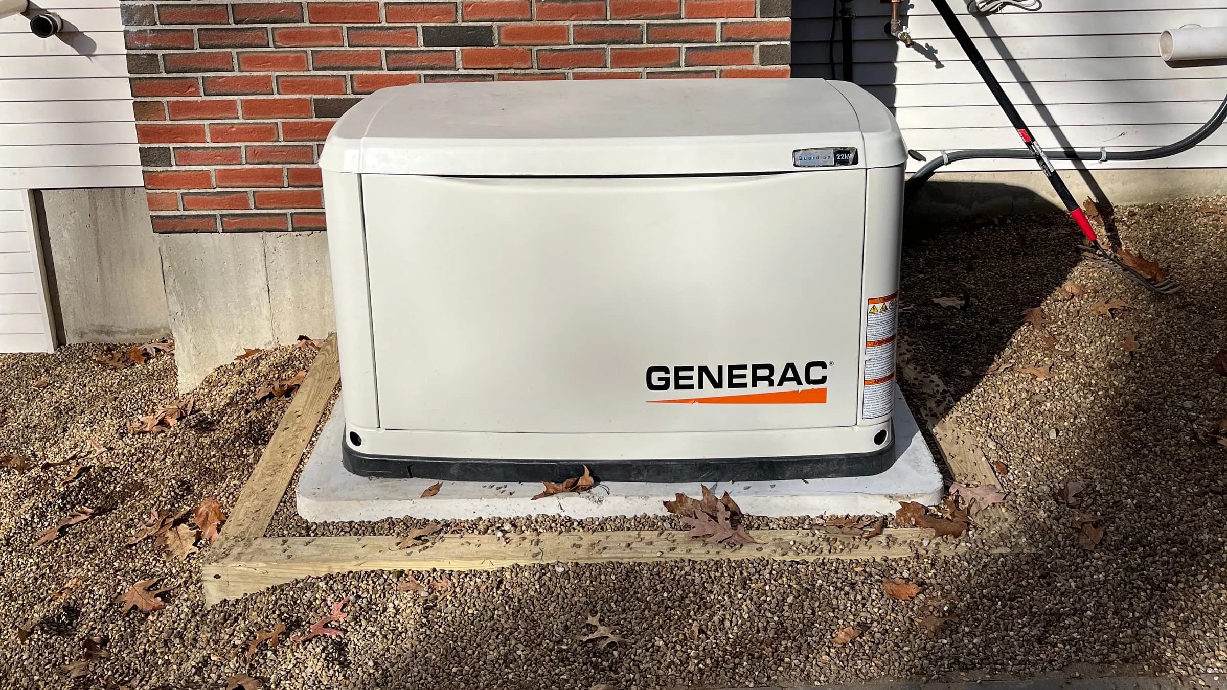 A Generac Guardian 22kW home standby generator on a concrete pad inside a timber-framed gravel bed, against a brick and vinyl-sided house on a sunny autumn day with fallen oak leaves.