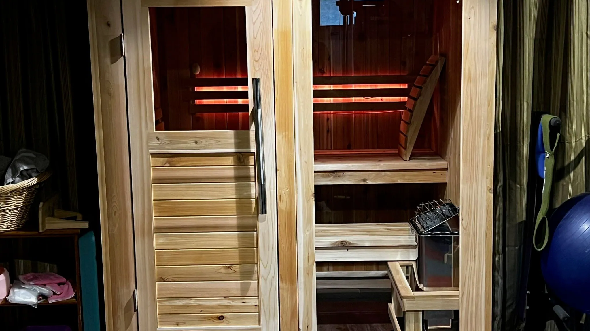 A cedar indoor sauna with a glass-panel door, red LED strip lighting behind the benches and an electric heater with stones, set in a home gym corner.