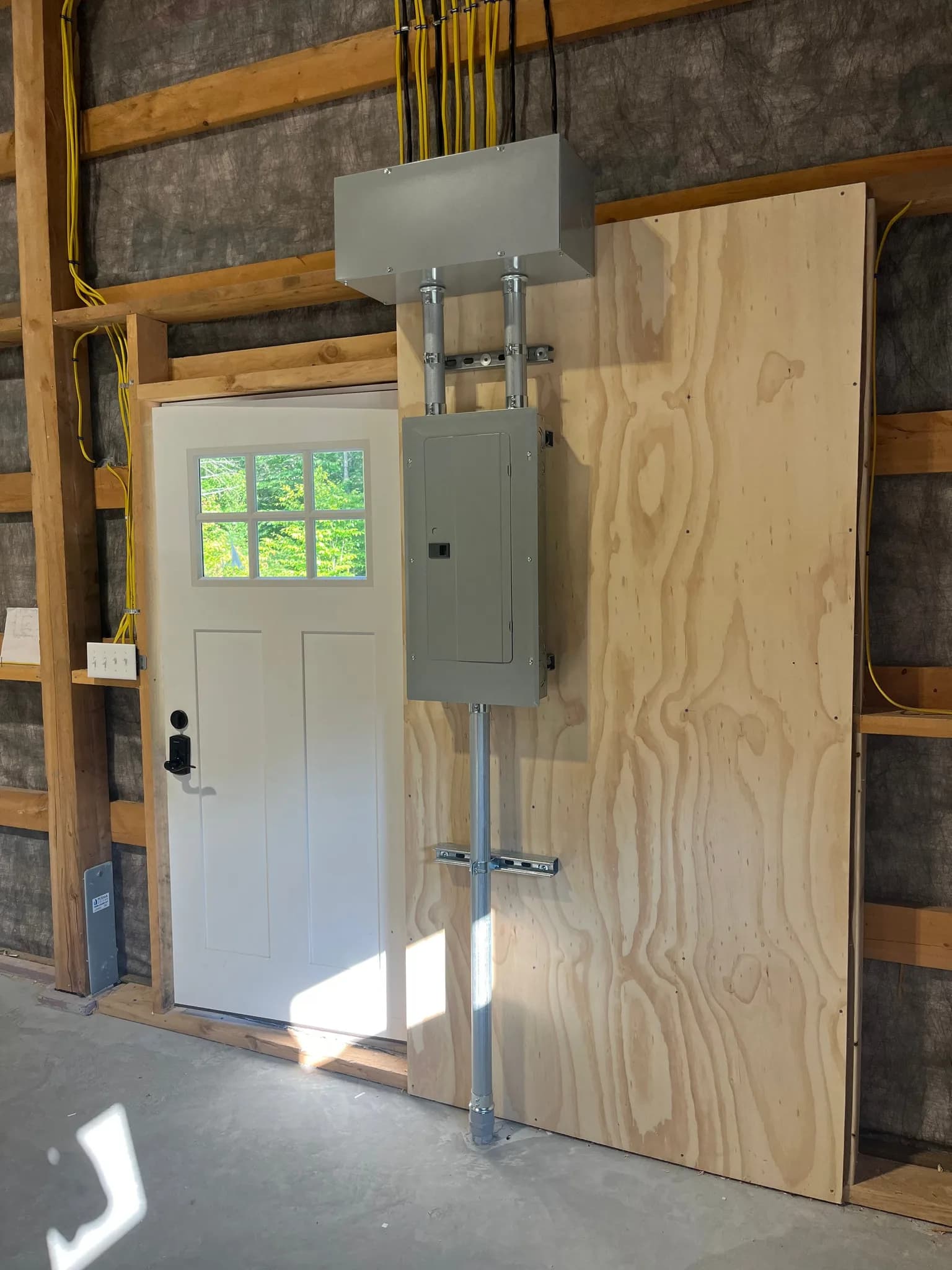 A new gray breaker panel on a plywood backboard in a framed garage, fed by two conduits from a junction box above, with a conduit run down to the slab beside a white entry door.