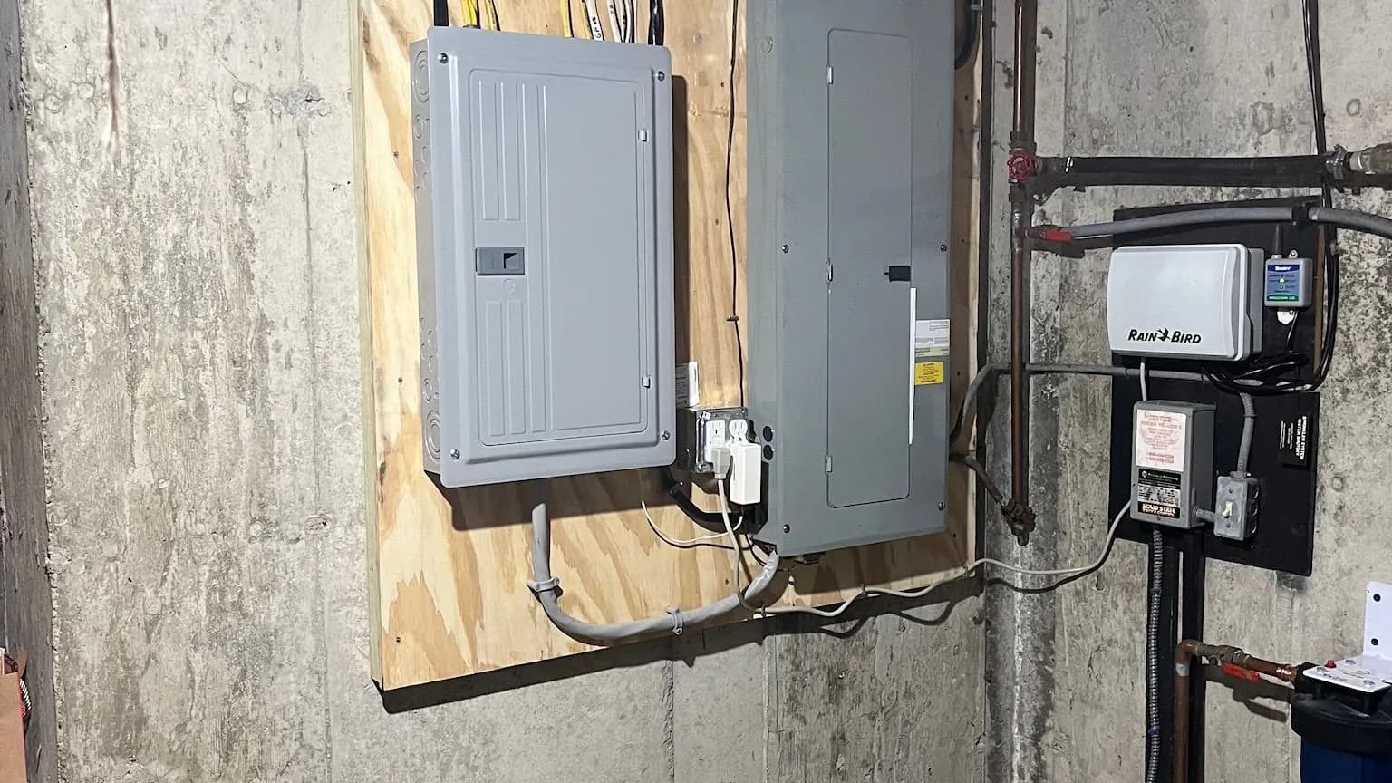 Two gray breaker panels mounted on a plywood backboard against a poured concrete basement wall, next to a Rain Bird irrigation controller, a pump control box and copper water pipes.