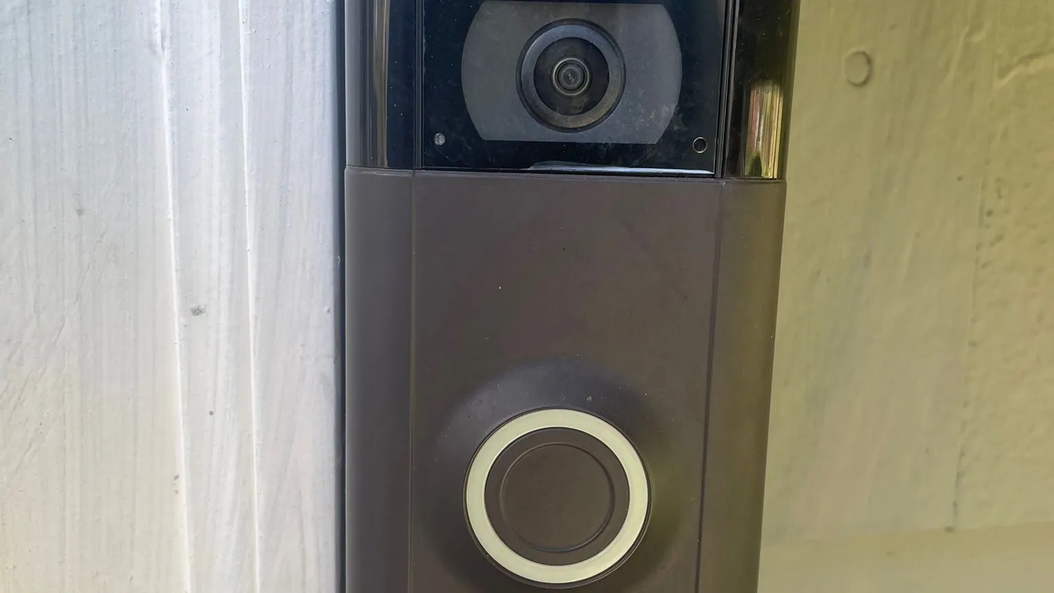 Close-up of a bronze video doorbell with a wide-angle camera lens and a lit ring button, mounted on white-painted door trim.