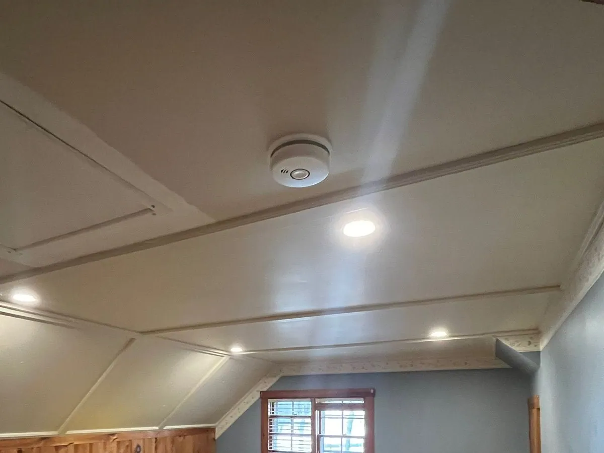 A ceiling-mounted smoke alarm and recessed LED lights in a white paneled ceiling with trim strips, in a room with blue walls, knotty pine wainscoting and a window.