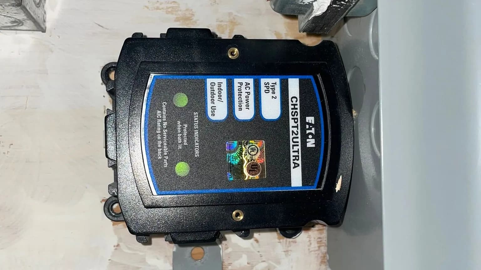 An Eaton CHSPT2ULTRA Type 2 whole-home surge protector with two green status lights lit, mounted on its side on a painted plywood backboard beside a gray electrical panel.