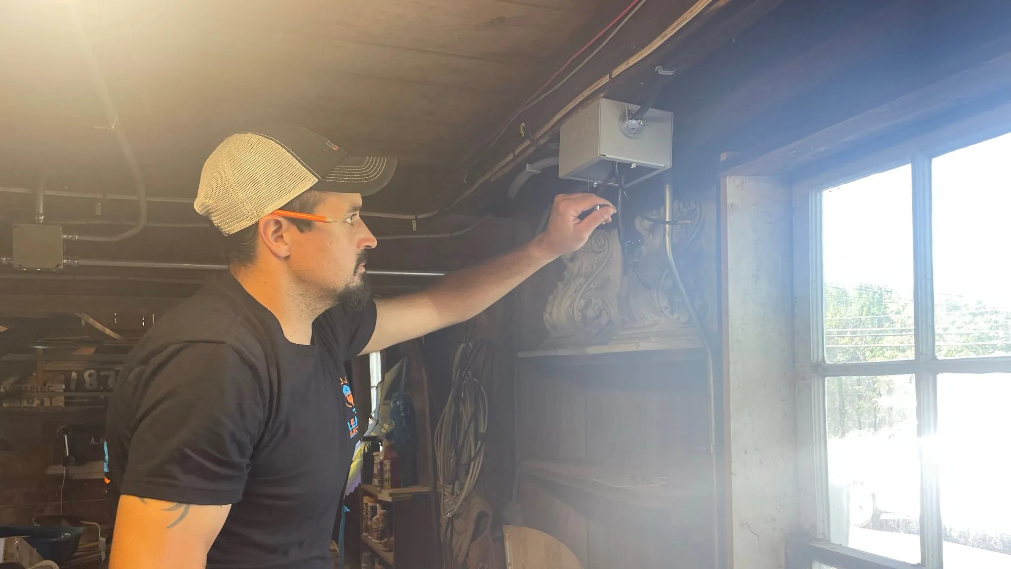 An electrician in a trucker cap and branded black T-shirt reaches up to wire a gray junction box under the beams of an old wooden workshop, with bright sun through the window.