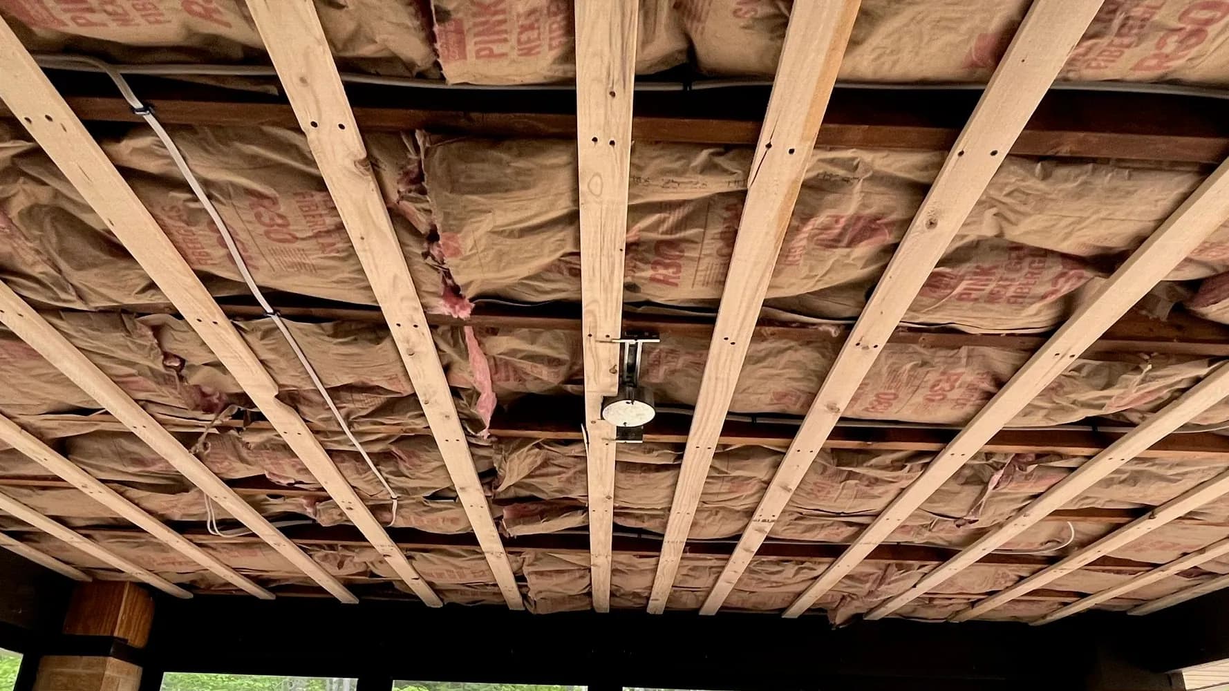 Wood furring strips run across a ceiling of paper-faced pink R30 fiberglass insulation, with a single light fixture box and cable runs roughed in, open to green trees at the far end.