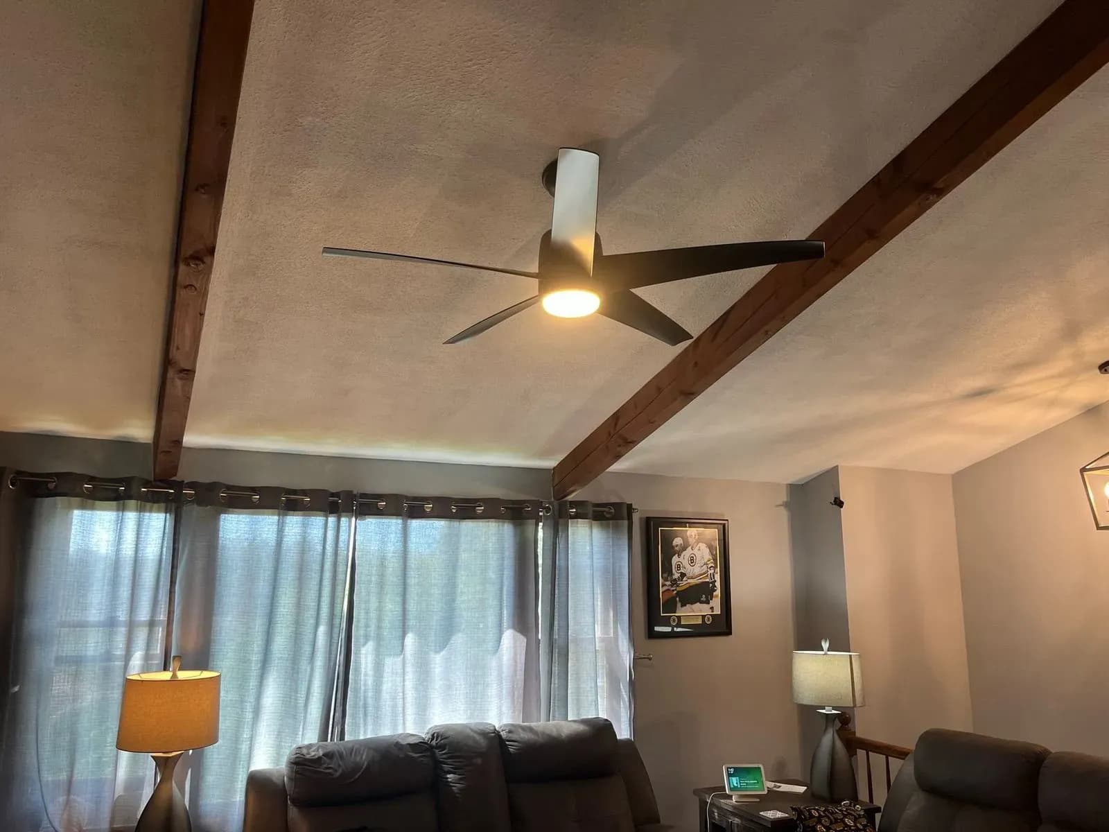 A black modern ceiling fan with an LED light centered on a sloped living room ceiling between two dark wood beams, sheer curtains and a leather sofa below.