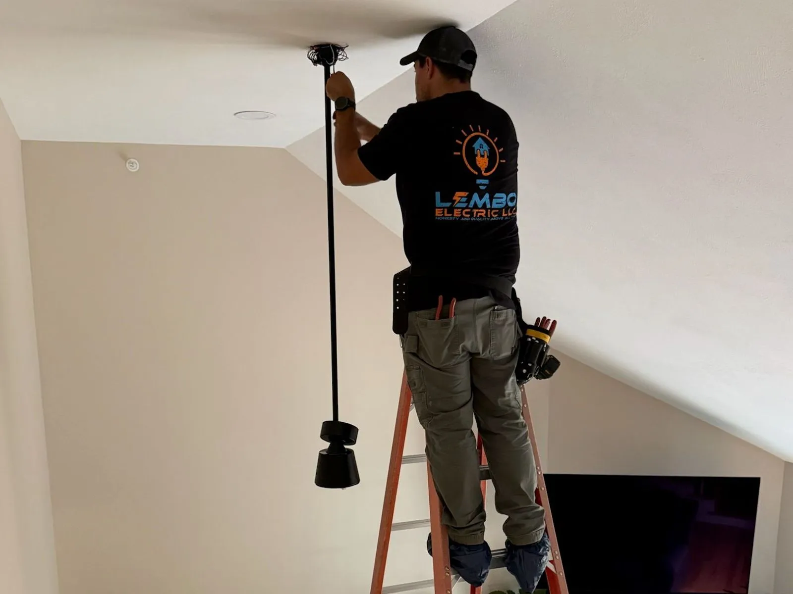 A Lembo electrician on an orange stepladder mounts a long ceiling fan downrod to a vaulted ceiling above a living room.