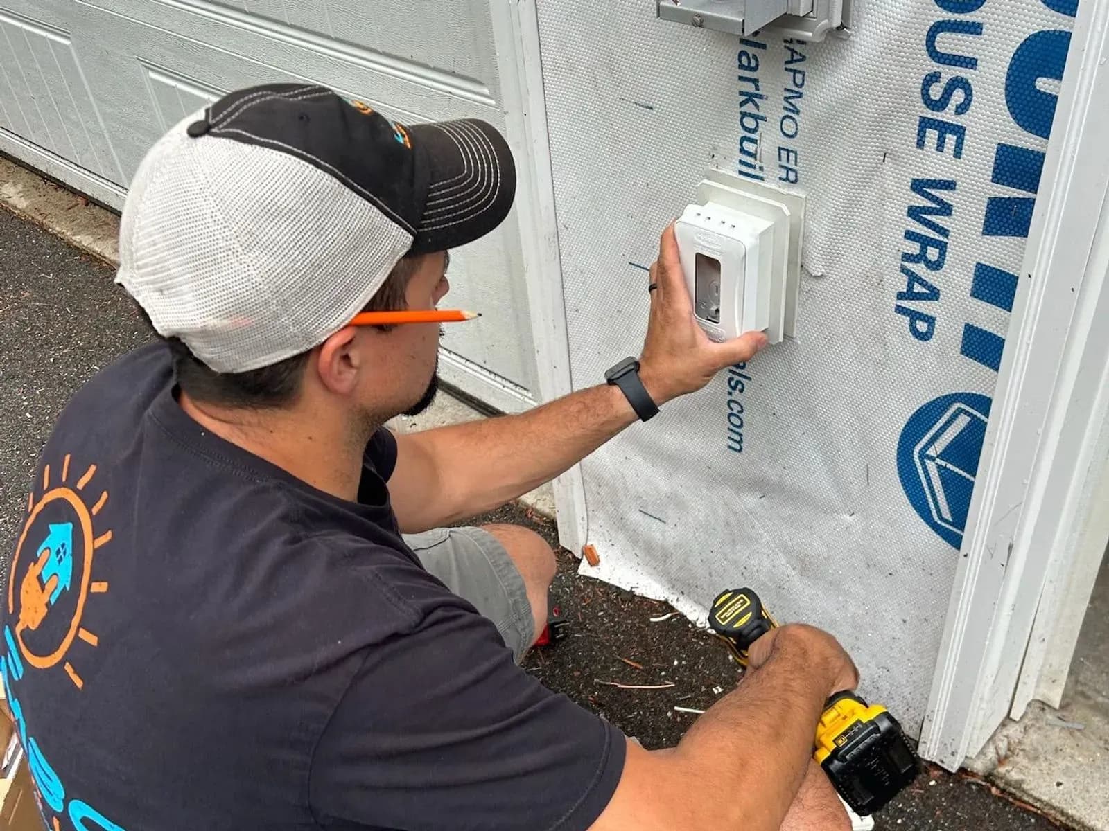 A Lembo Electric electrician in a cap crouches by a garage door, pressing a new white weatherproof outlet box onto the house wrap, a drill in the other hand.