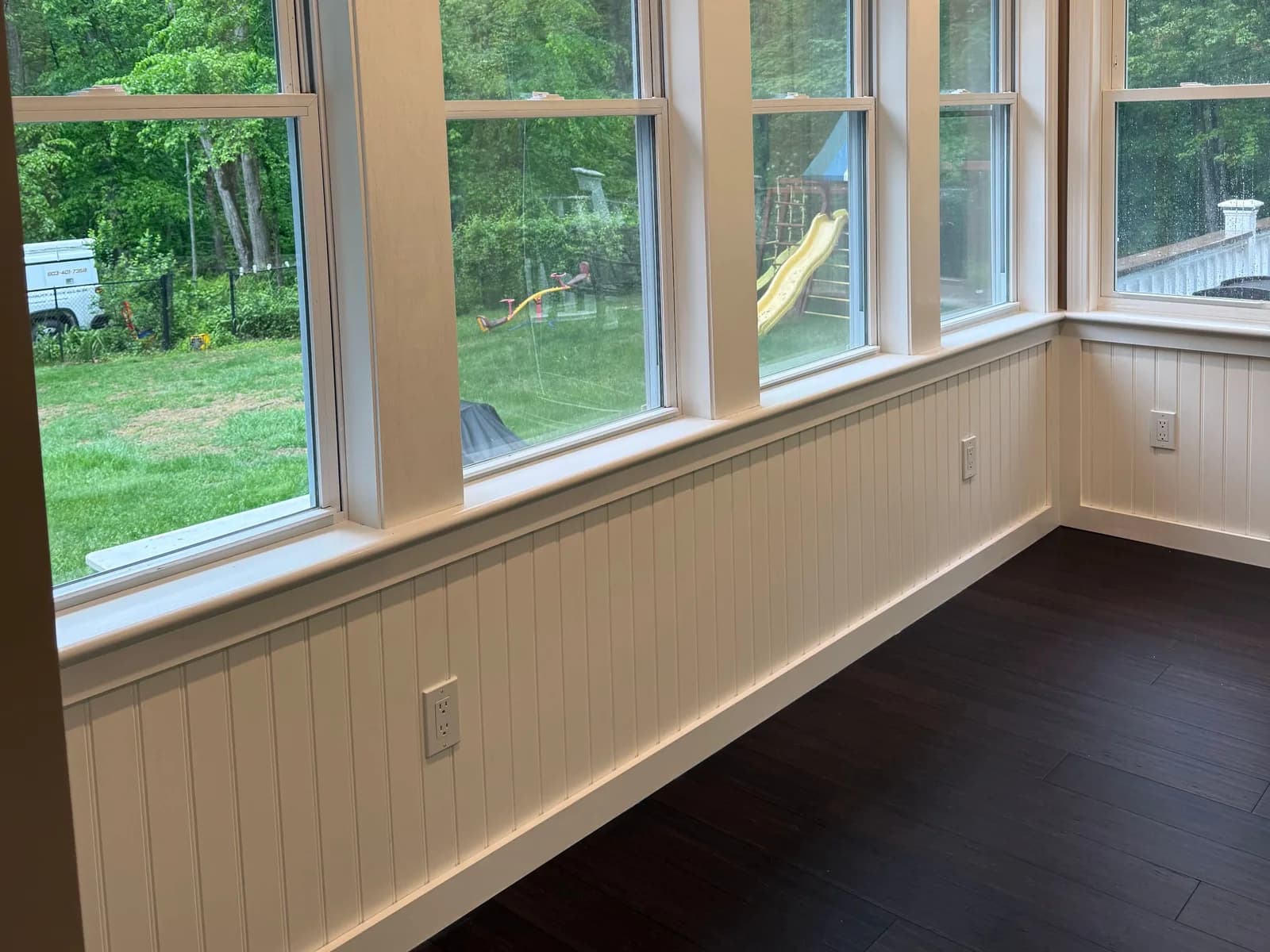 A new sunroom with a wall of double-hung windows over white beadboard wainscot, new duplex outlets set in the paneling along the wall, a vaulted beadboard ceiling and a dark wood floor.