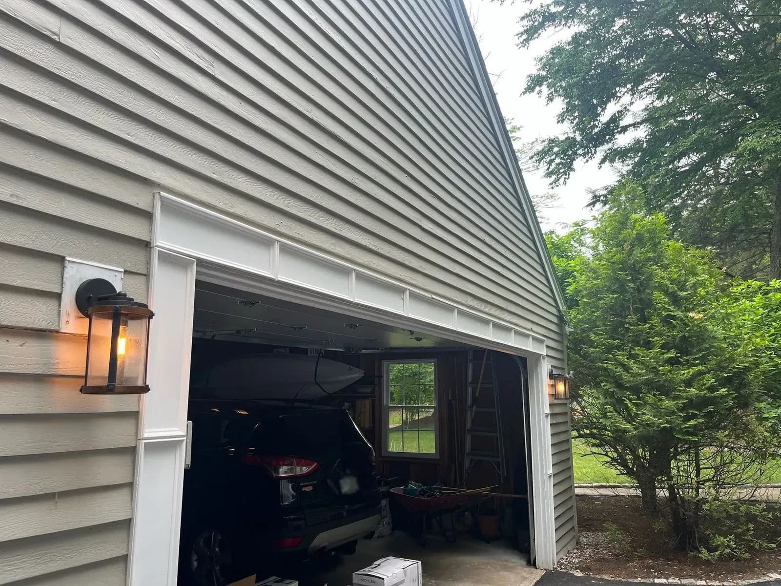 Black lantern wall lights glowing on both sides of an open garage door on a gray-sided home, an SUV and a kayak inside the garage and green trees beside it.