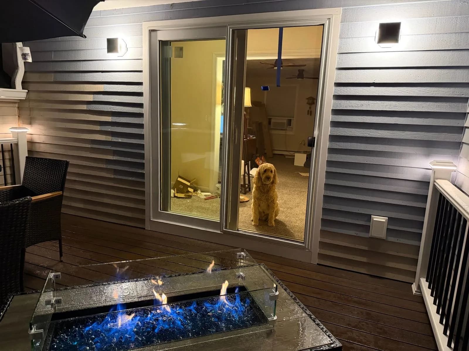 A deck at night with two modern wall lights washing the siding beside a sliding door, a fire table with blue glass flames in front and a dog looking out.