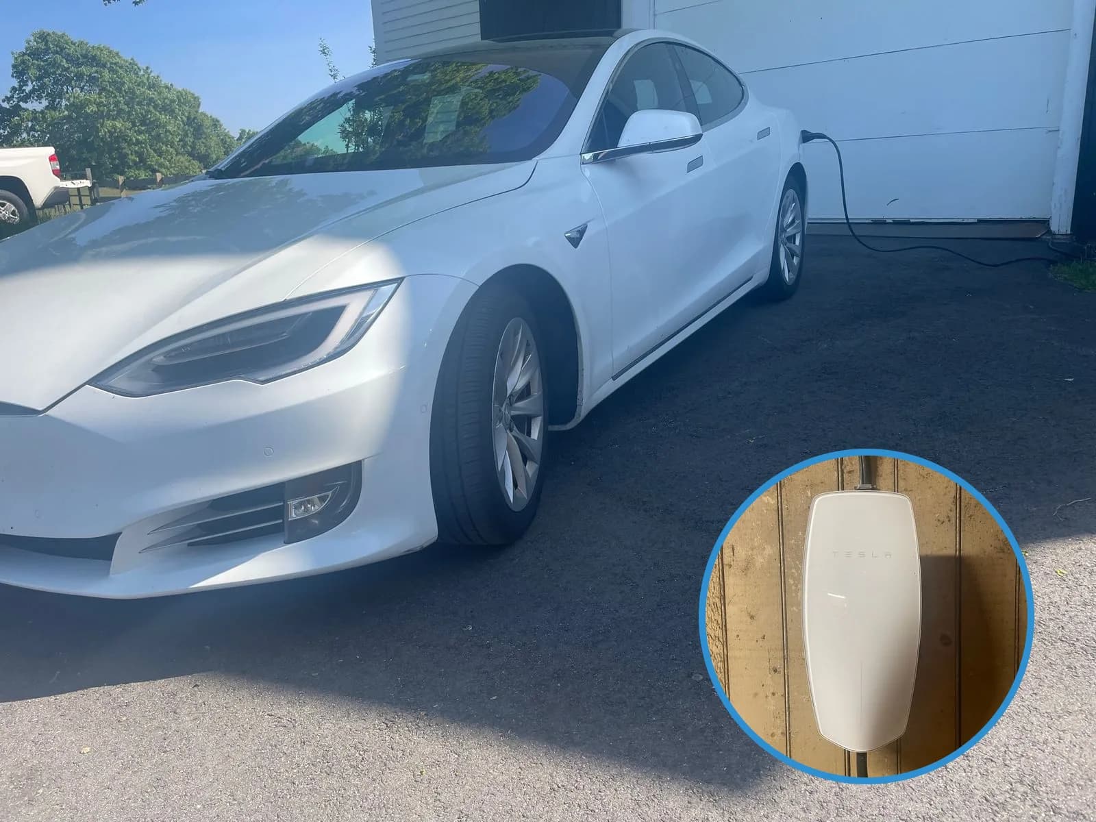 A white Tesla sedan parked in a driveway with its charging cable plugged in at the rear and running back to the garage, and a round inset of the white Tesla Wall Connector on a weathered beadboard wall.