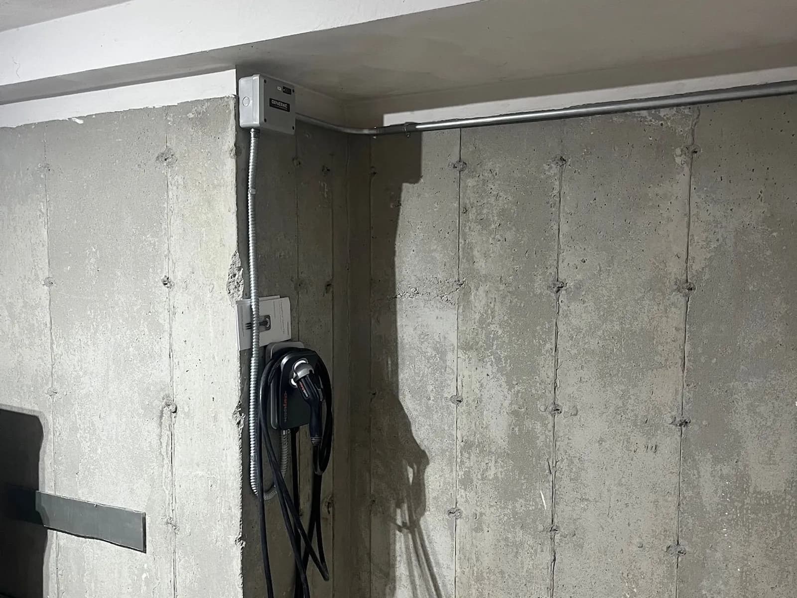 A ChargePoint home EV charger with its coiled cable on a board-formed concrete garage wall, fed by flexible metal conduit from a gray Generac box at the ceiling, where a steel conduit run follows the beam across the wall.