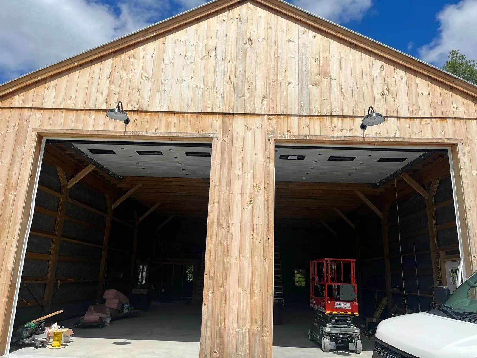 The board-and-batten front of a new pole barn with two gooseneck barn lights over its open overhead doors.