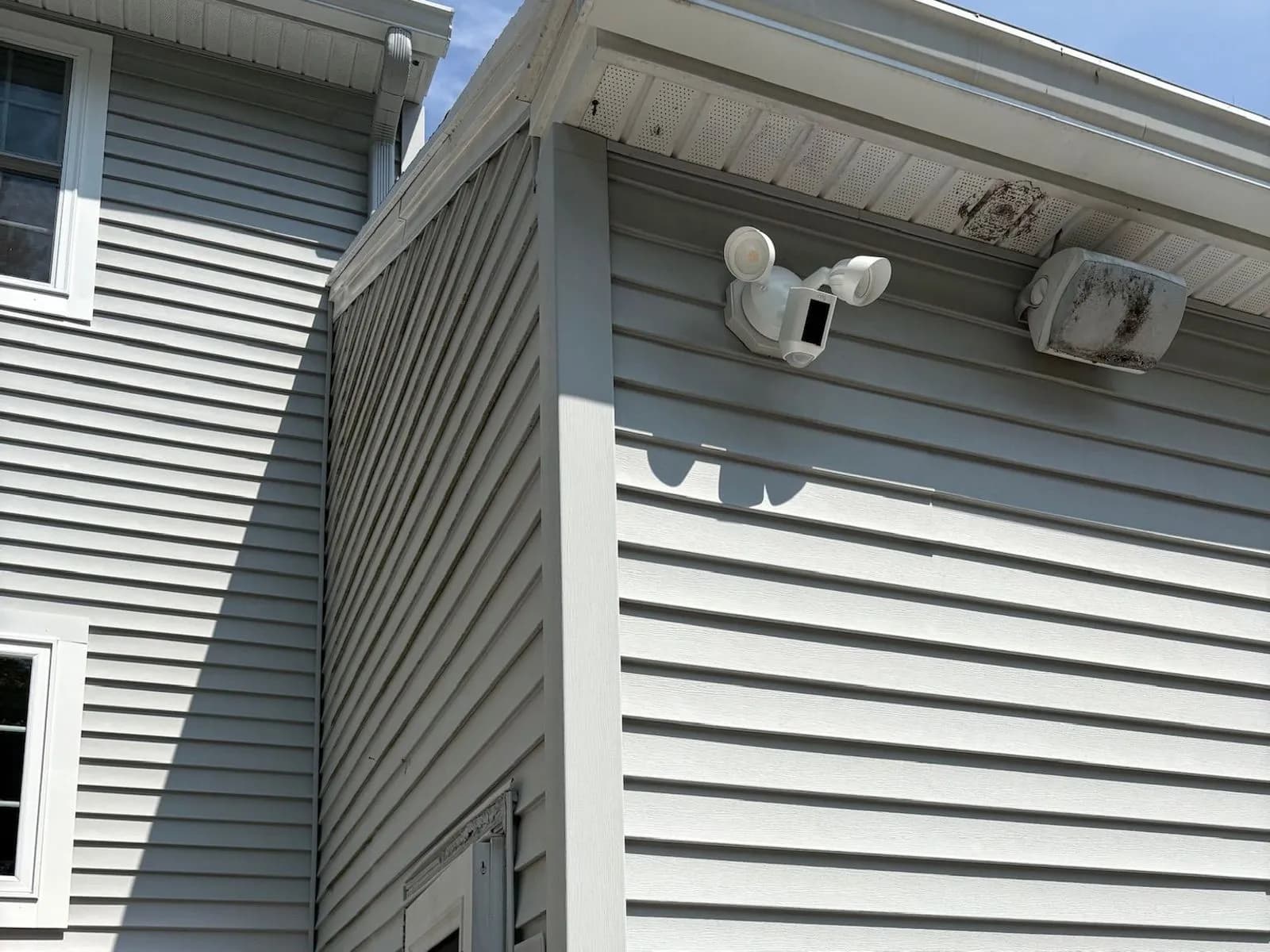 The corner of a gray sided house with a white floodlight security camera mounted under the eave.