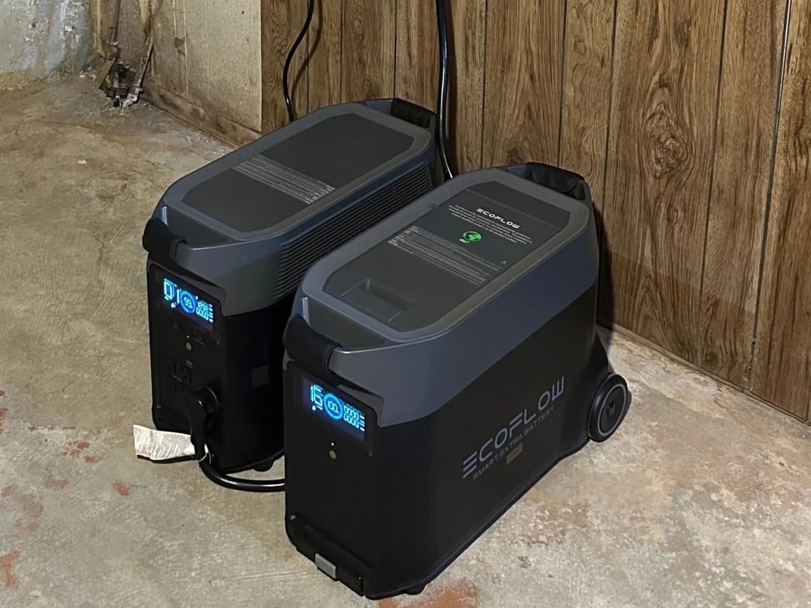Two black EcoFlow battery units side by side on a basement floor against wood paneling, their blue displays lit, the power cable running up the wall from behind them.