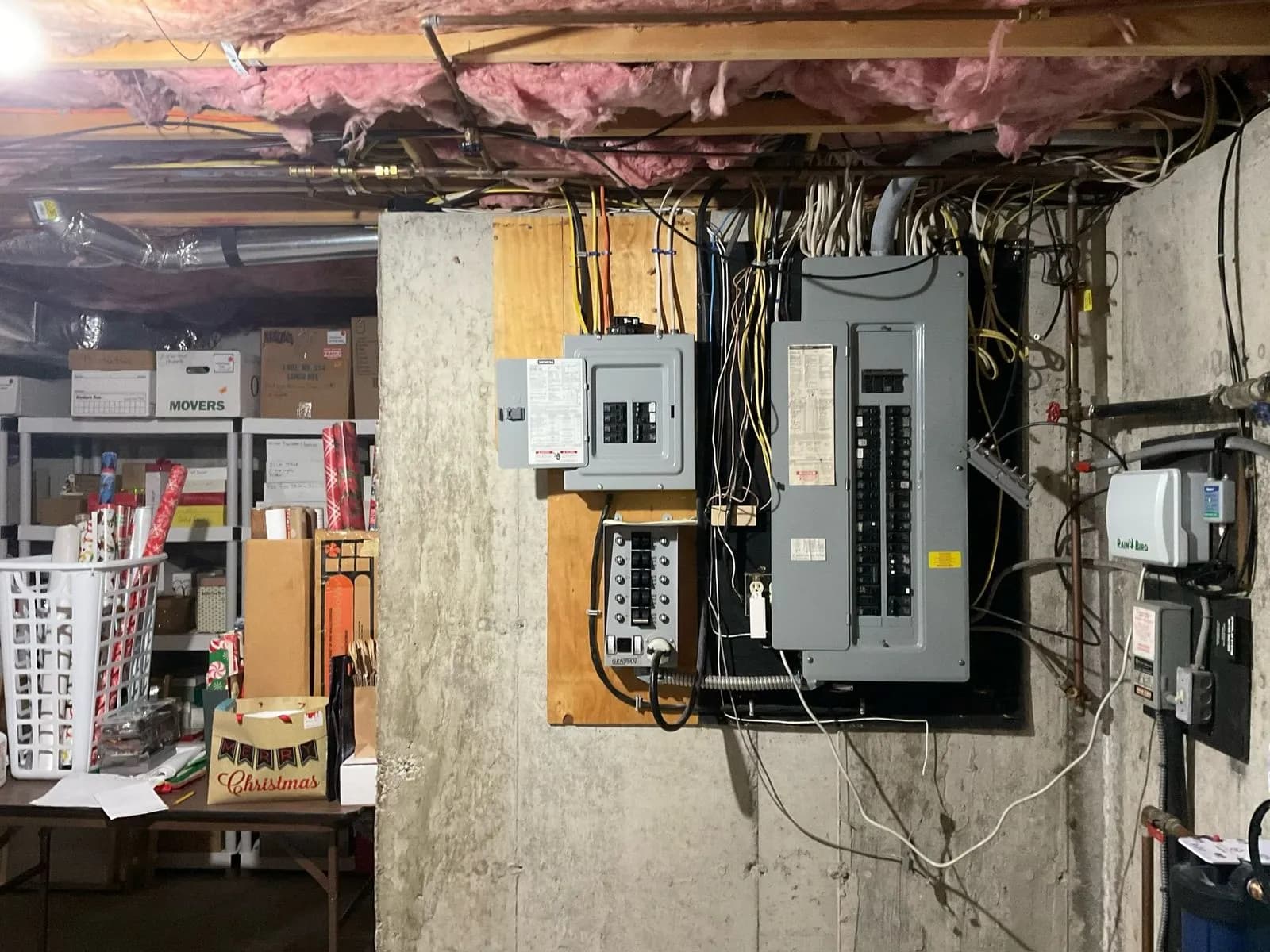 An unfinished basement wall with a main breaker panel, a smaller transfer panel beside it, and bundled wiring running up into the joists, shelving and boxes in the room behind.