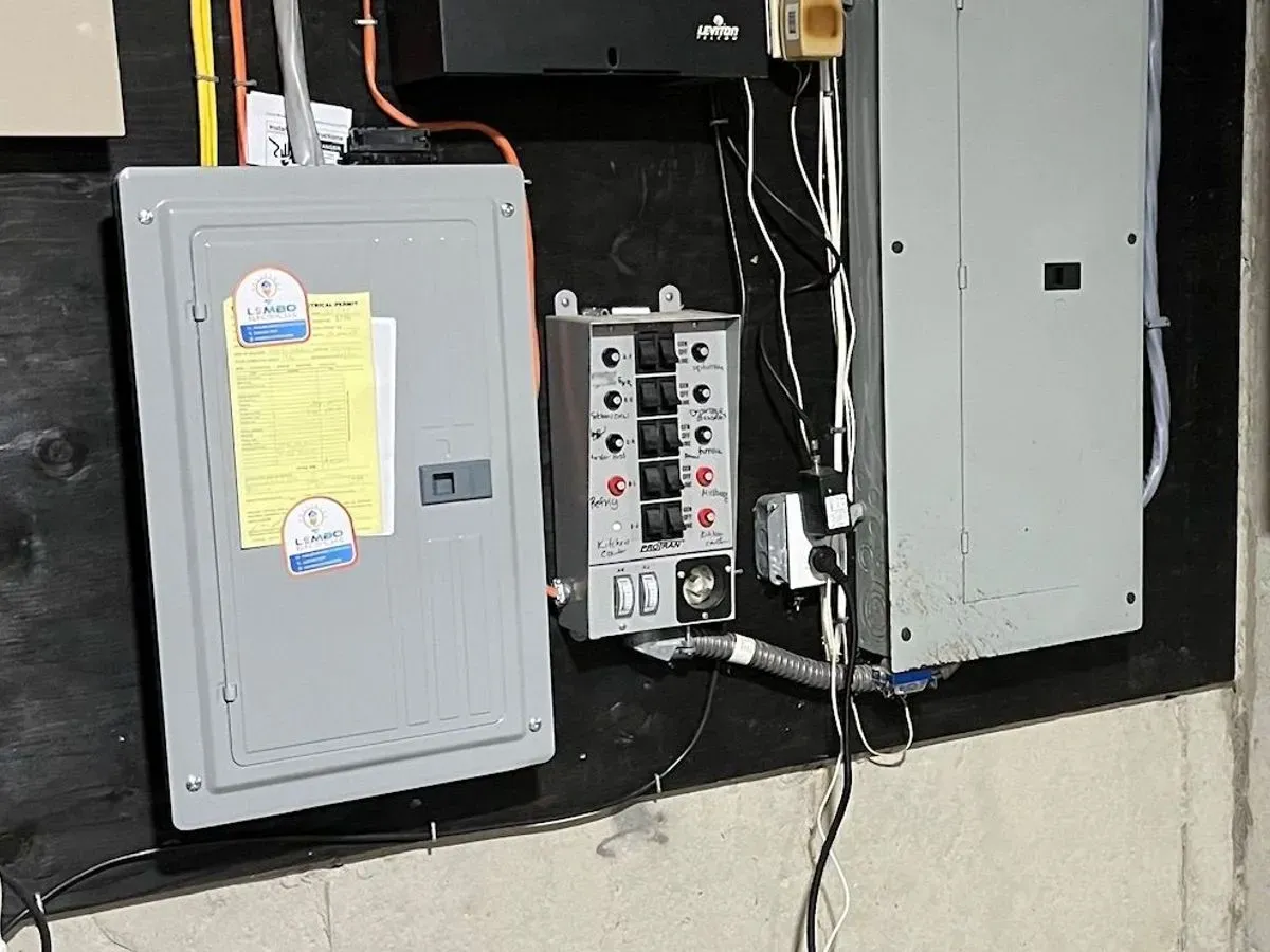 A breaker panel with Lembo Electric inspection stickers on a black backboard in a basement, a manual transfer switch with a row of labeled circuit switches mounted beside it.