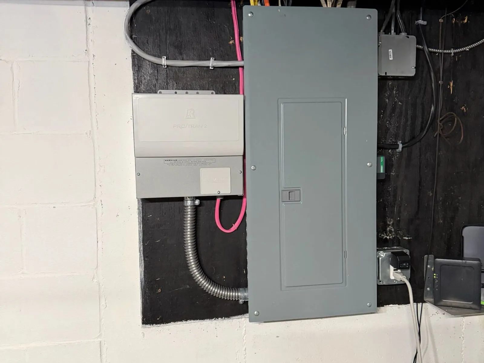 A gray breaker panel on a black backboard set into a white-painted block basement wall, flexible conduit looping from a smaller box beside it.
