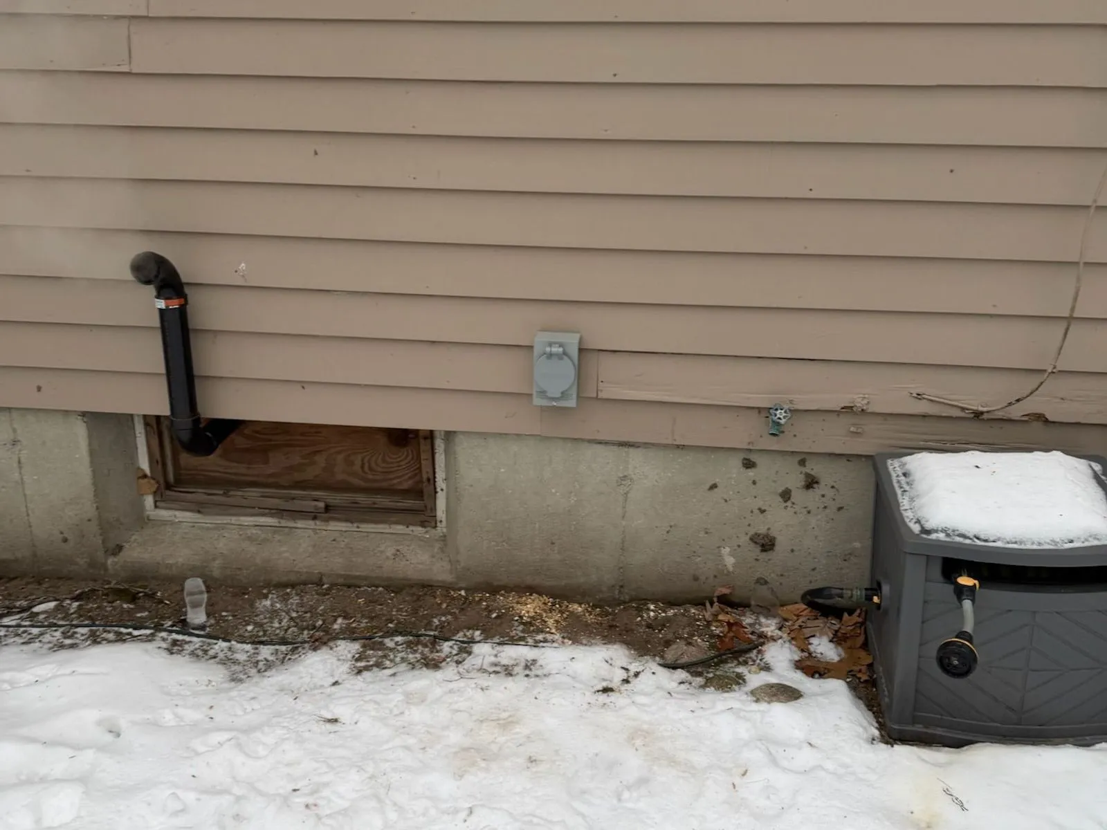 A gray weatherproof power inlet on tan clapboard siding above a snowy yard, beside a black vent pipe and a basement window well.