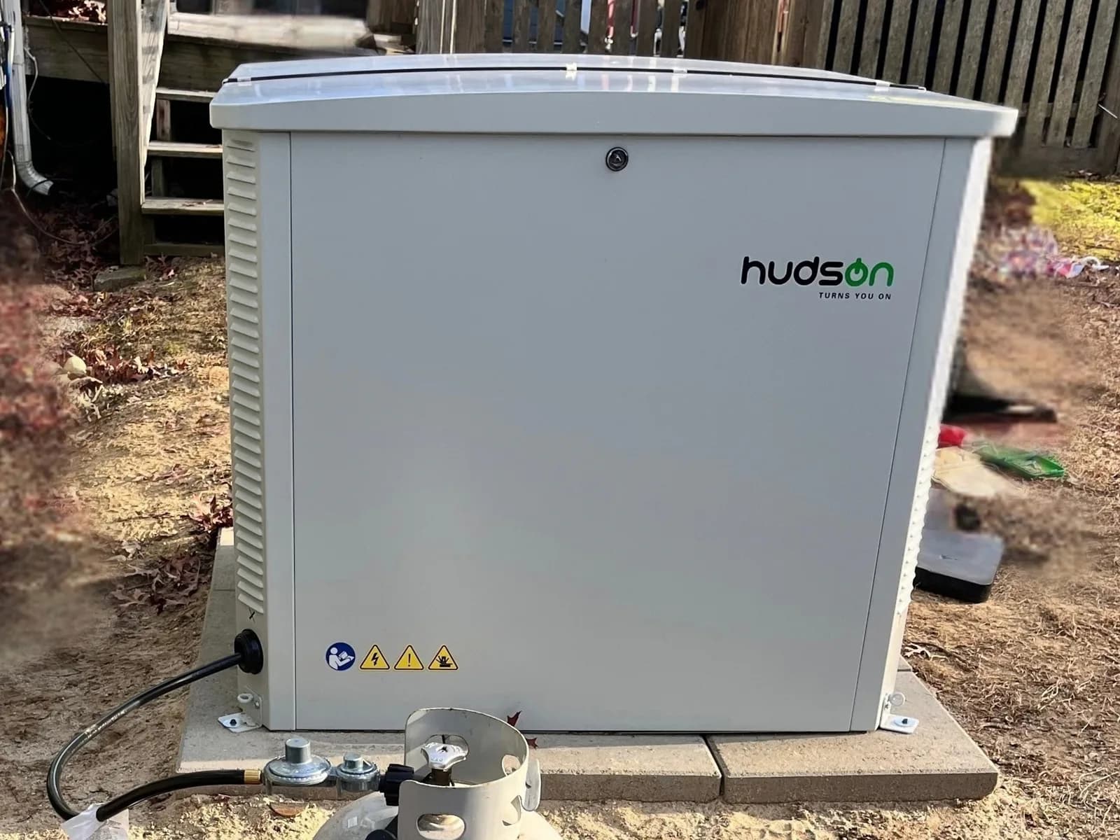 Close view of a white Hudson generator enclosure with vented sides and a locking lid, set level on square pavers, a black propane hose and regulator connected at its lower corner.