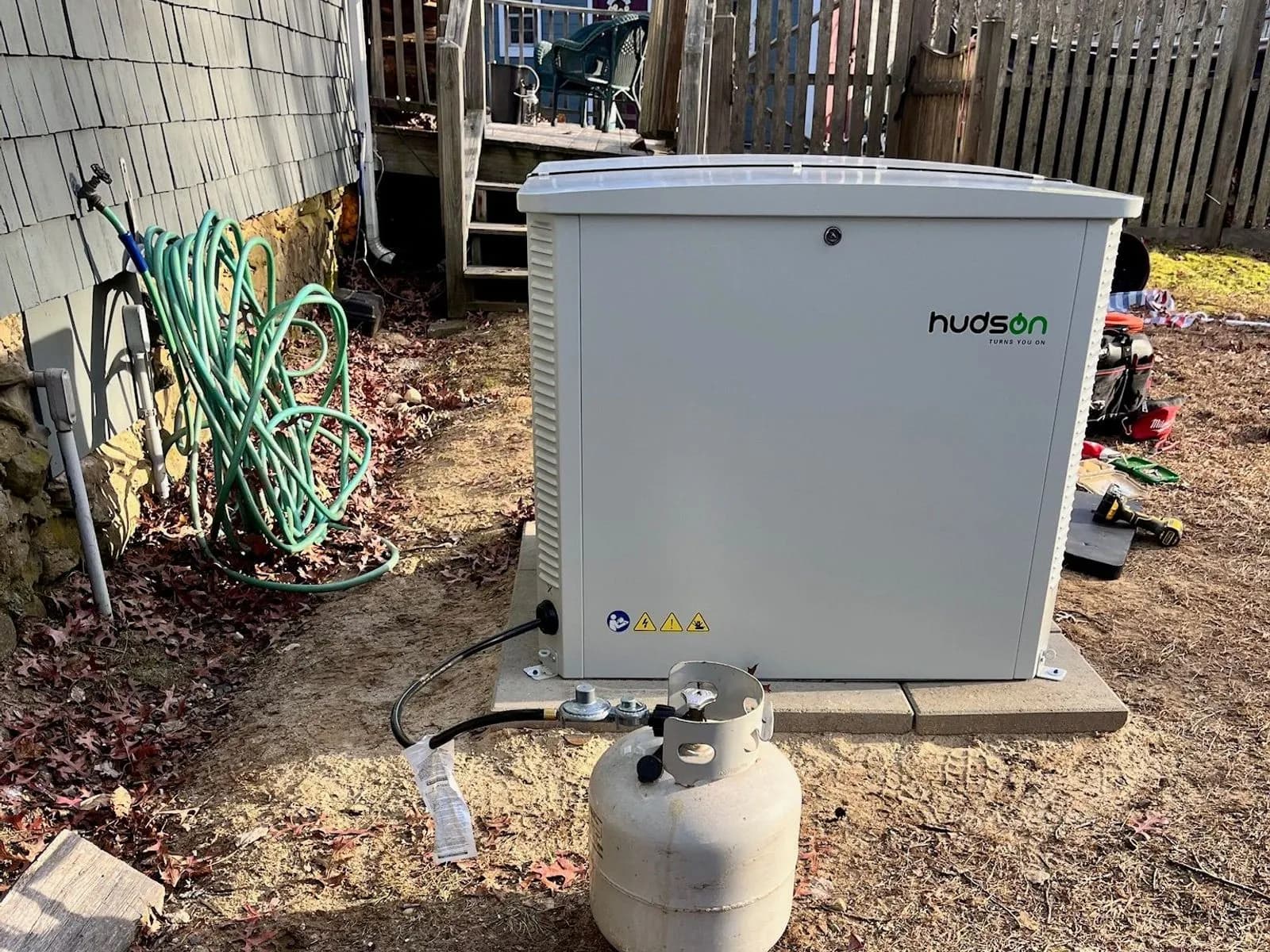 A white Hudson generator enclosure on a paver pad in a leafy side yard beside a shingled house, a propane tank connected to it by a black hose, with a coiled garden hose on the wall and a wooden fence behind.