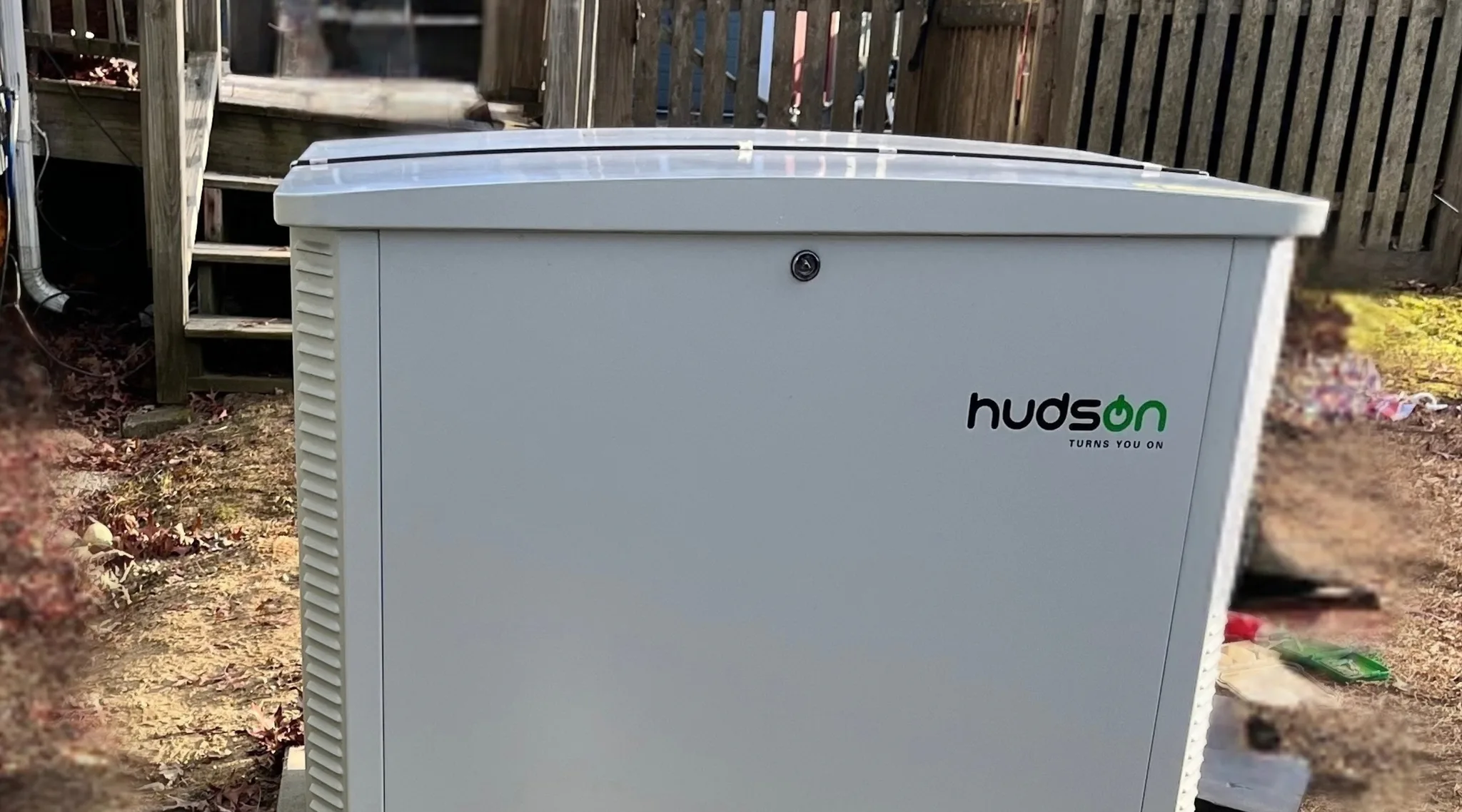 A white Hudson generator enclosure with vented sides and a locking lid standing in a leaf-strewn side yard, deck stairs and a weathered wooden fence behind it.
