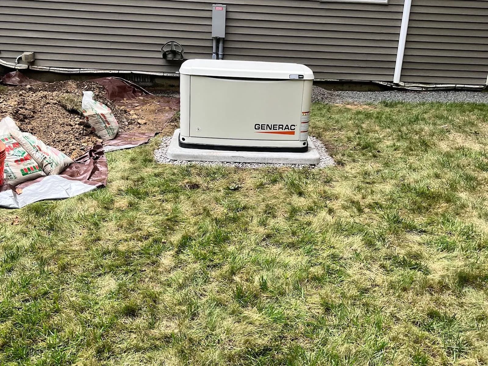 A white Generac standby generator on a pad in a green lawn beside a gray-sided house, a tarp and bags of materials at the edge of a fresh dig.