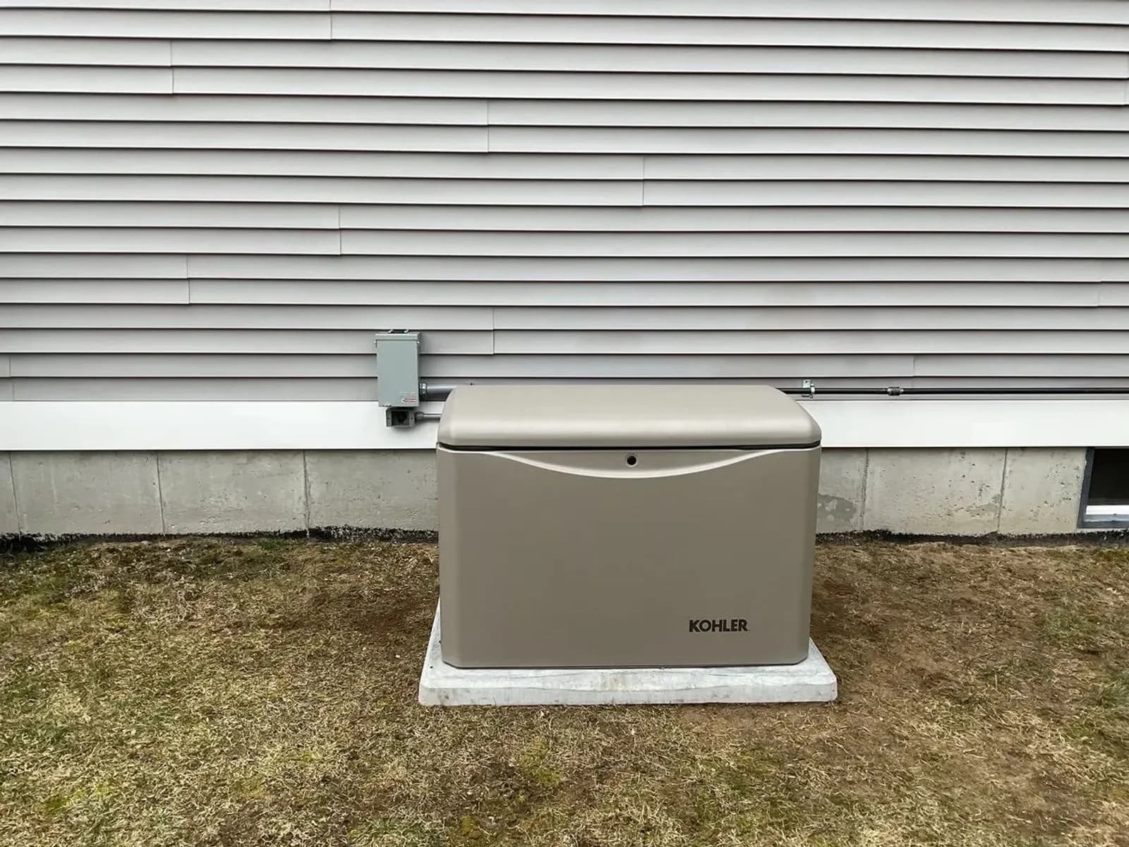 A tan Kohler standby generator on a white concrete pad against white vinyl siding, a gray disconnect on the wall above it and a trimmed lawn in front.