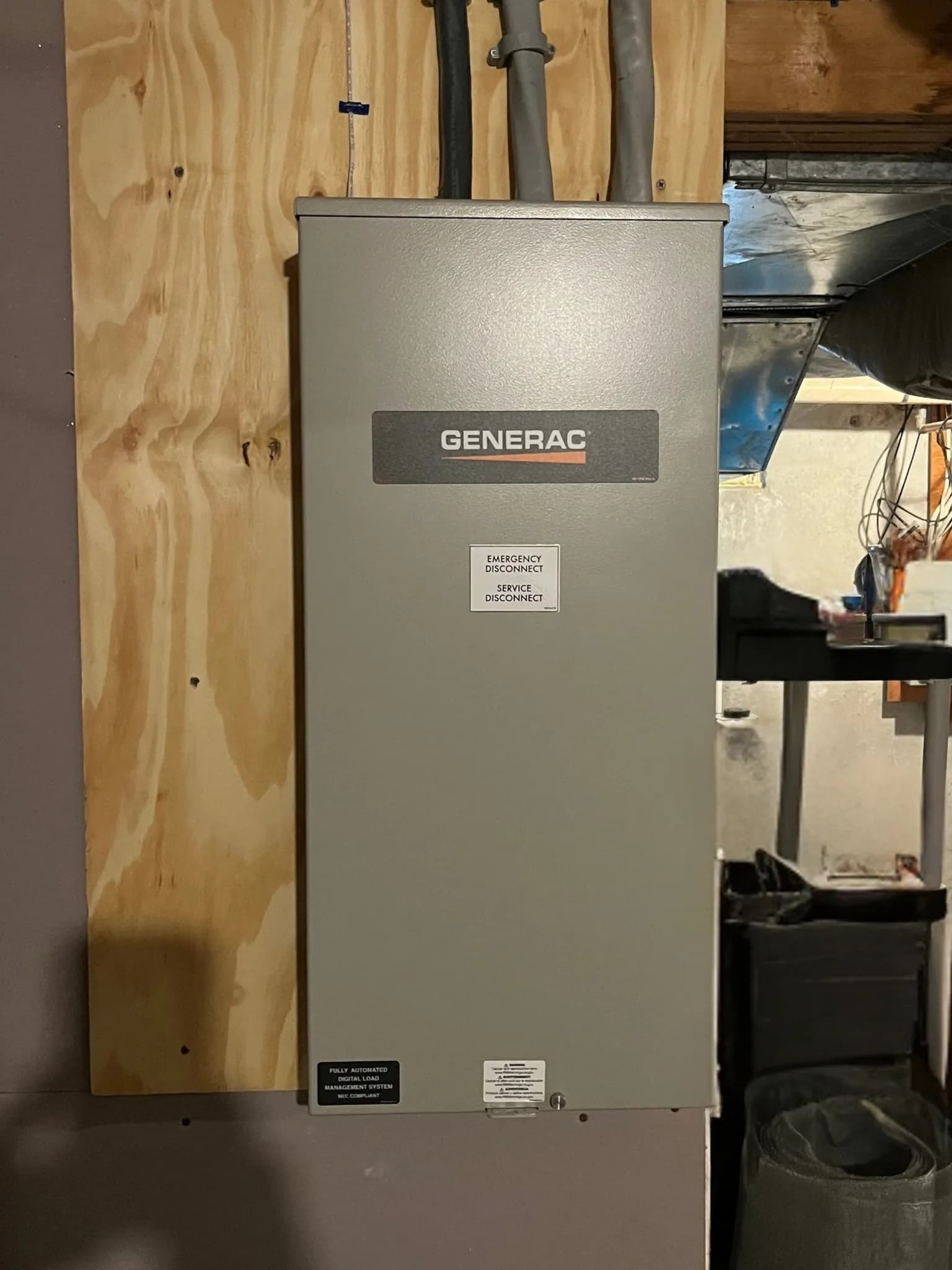 A gray Generac automatic transfer switch marked emergency service disconnect, mounted on a plywood backboard in a basement with three conduits running up into the ceiling.