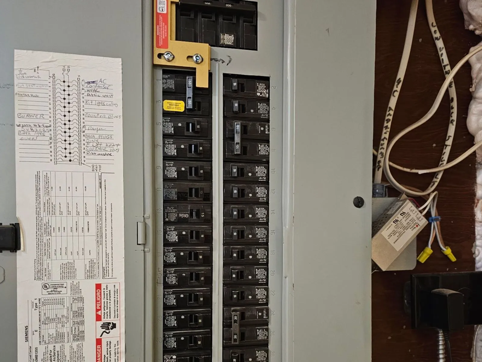 A breaker panel with a yellow sliding interlock plate over the main and generator breakers, a handwritten circuit directory on the open door beside it.