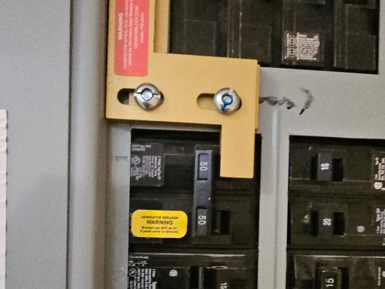 Close-up of a gold generator interlock plate bolted into a breaker panel above a 50-amp two-pole generator breaker with a yellow warning sticker.