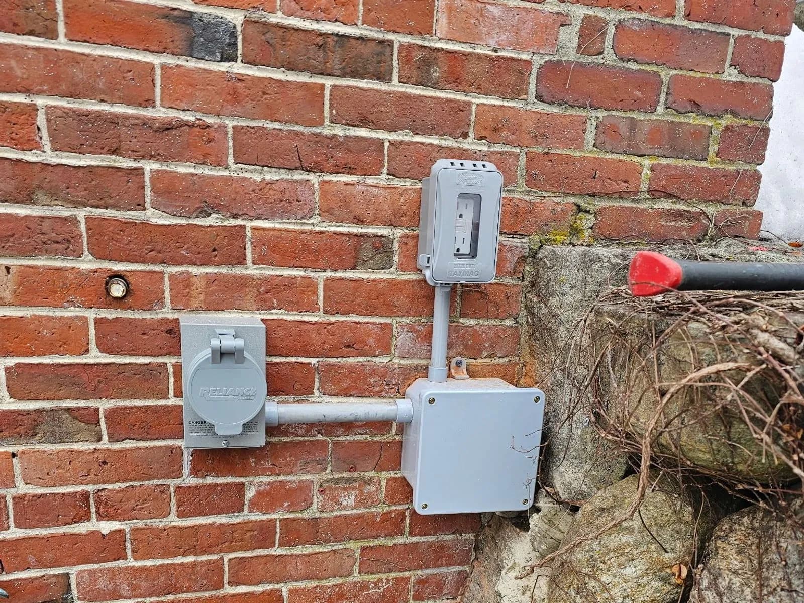 Two gray weatherproof boxes on a red brick wall joined by gray conduit, a power inlet with its hinged cover at left and a disconnect above a junction box, beside a fieldstone foundation.