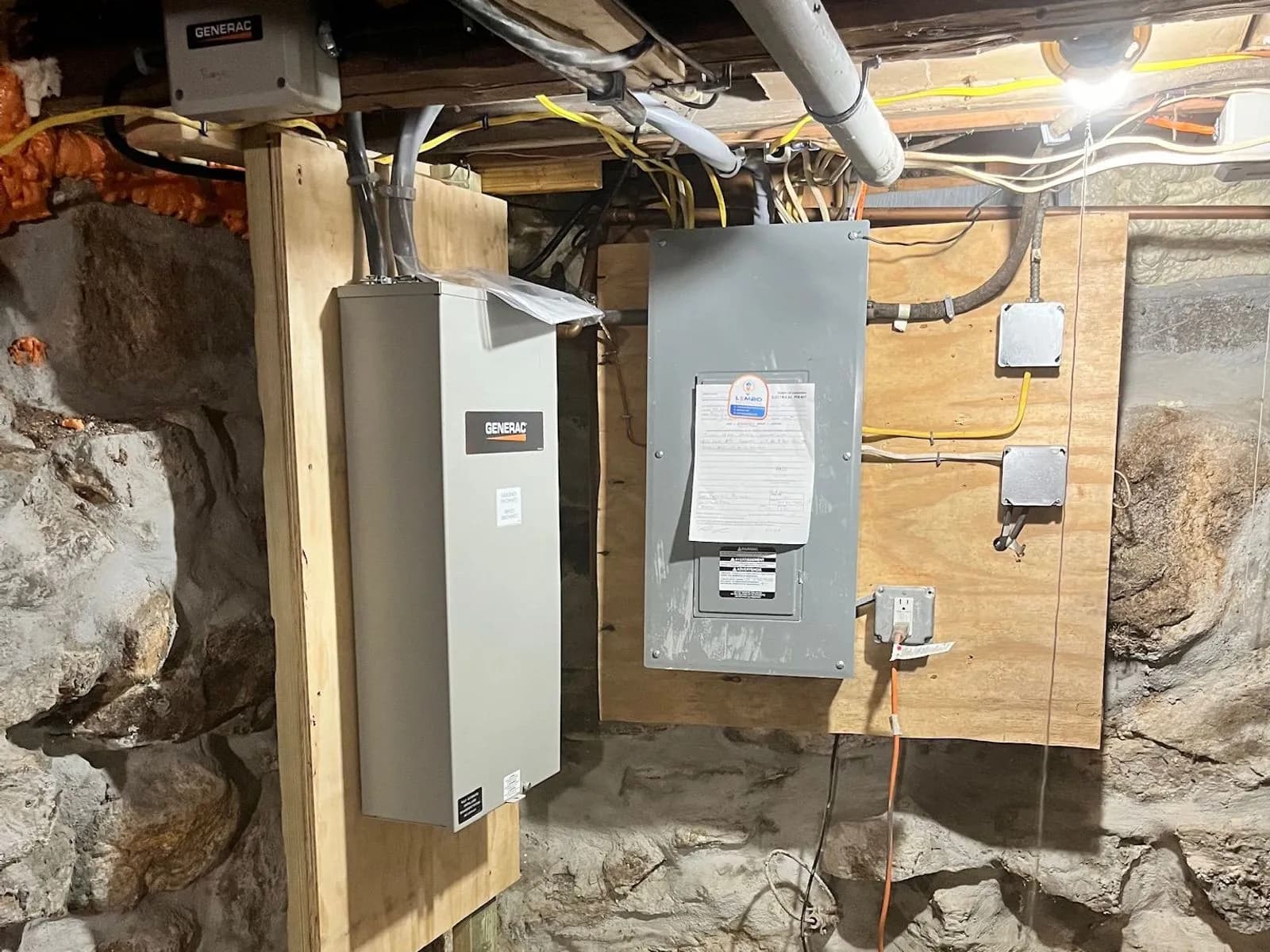 A basement with a fieldstone wall and sandy floor, a gray Generac transfer switch on a wooden post beside a panel on a plywood backboard, conduit running along the joists above.