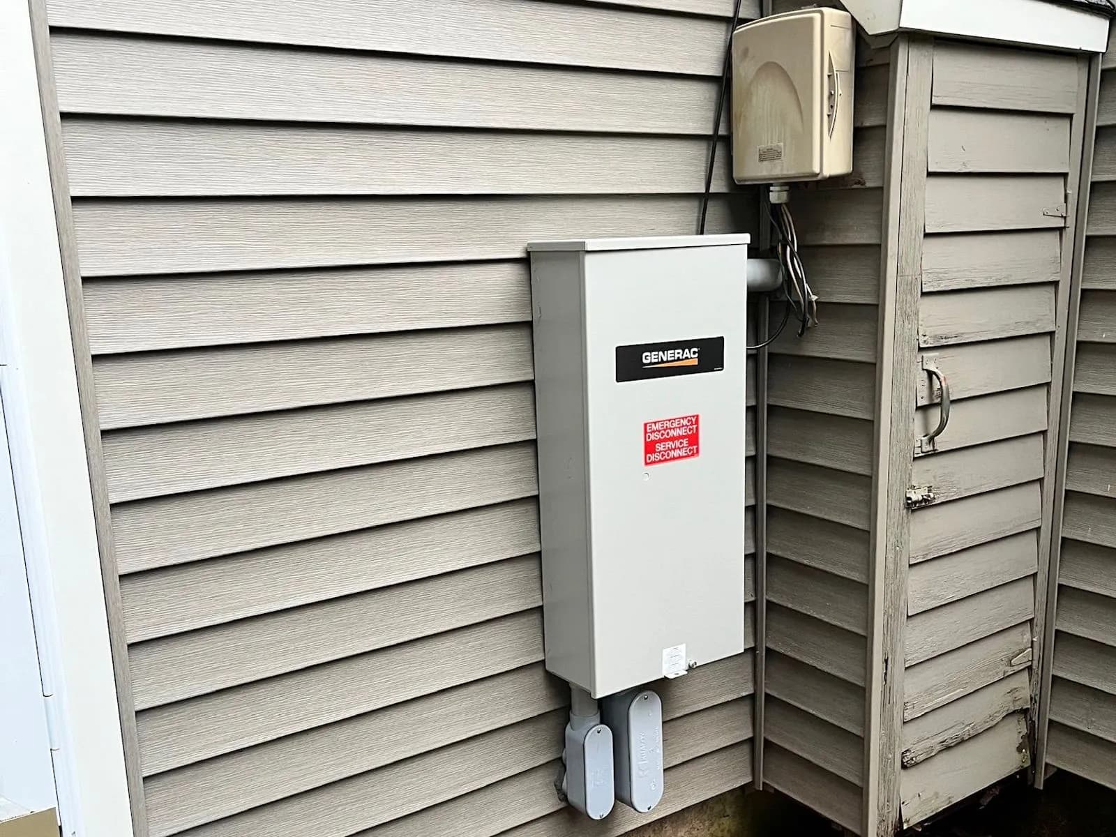 A gray Generac automatic transfer switch mounted on gray vinyl siding beside a small shed-roofed bump-out, conduit dropping from its base to the red mulch below.