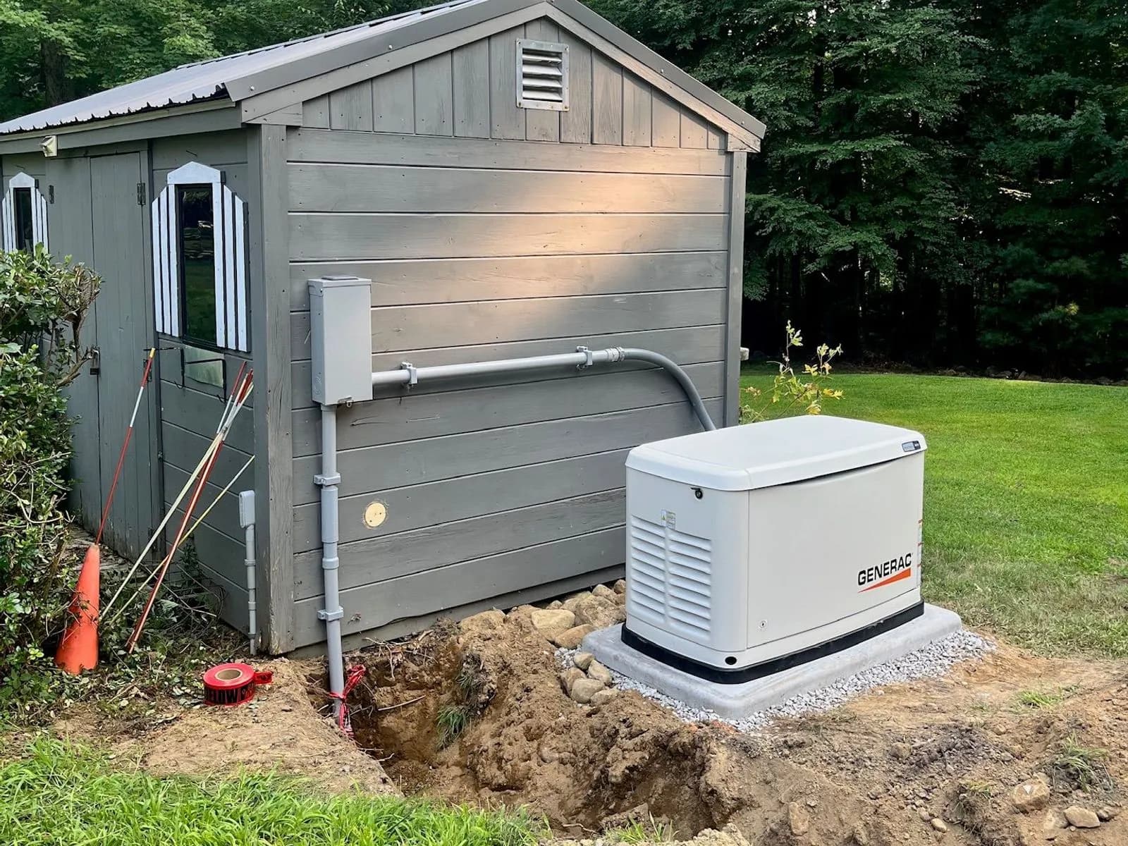 A Generac standby generator on a fresh pad beside a gray storage shed, conduit running along the shed wall to a disconnect, with the trench still open in the lawn.