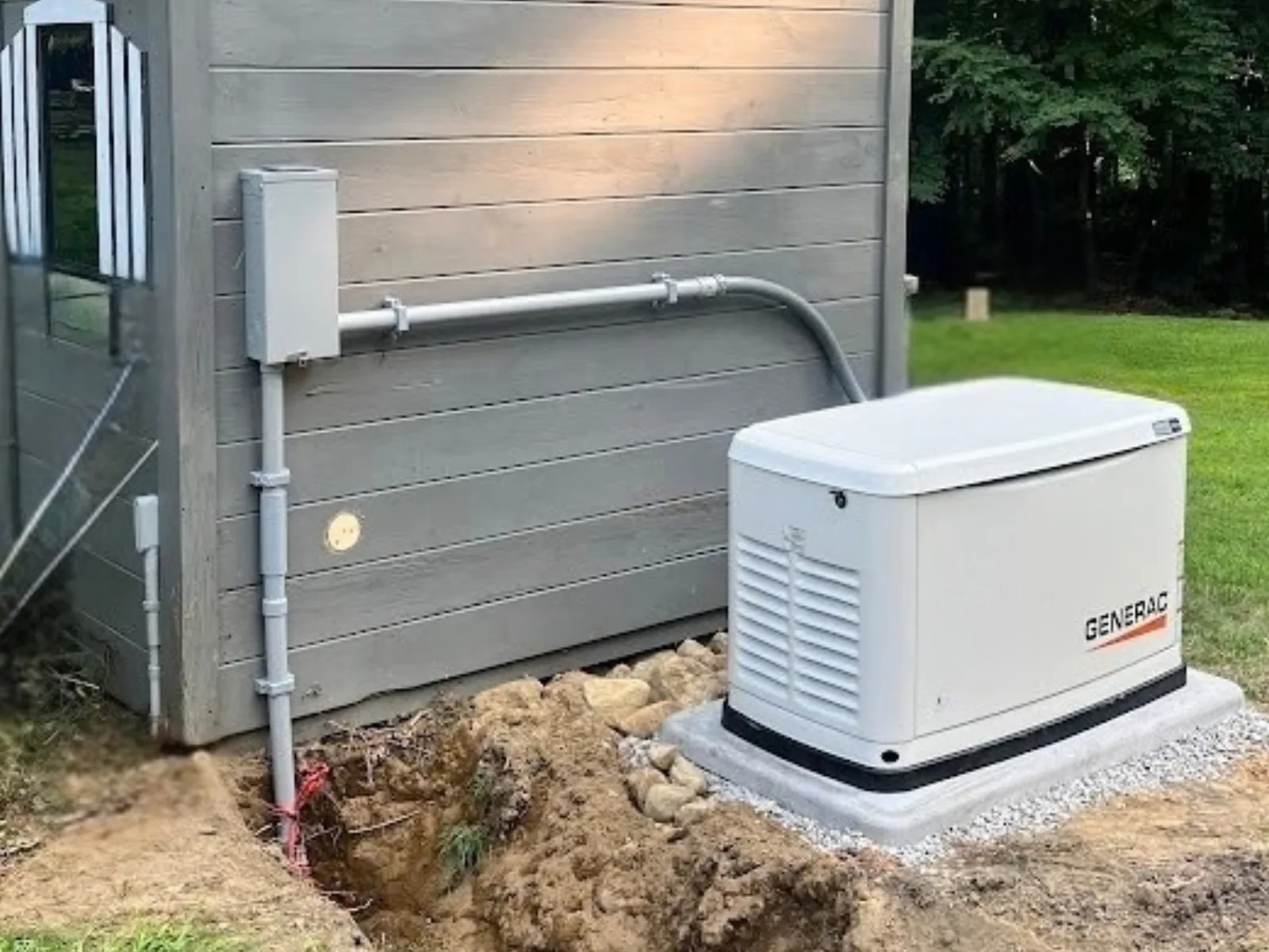 A Generac standby generator on a fresh pad beside a gray storage shed, conduit running along the shed wall to a disconnect, with the trench still open in the soil.