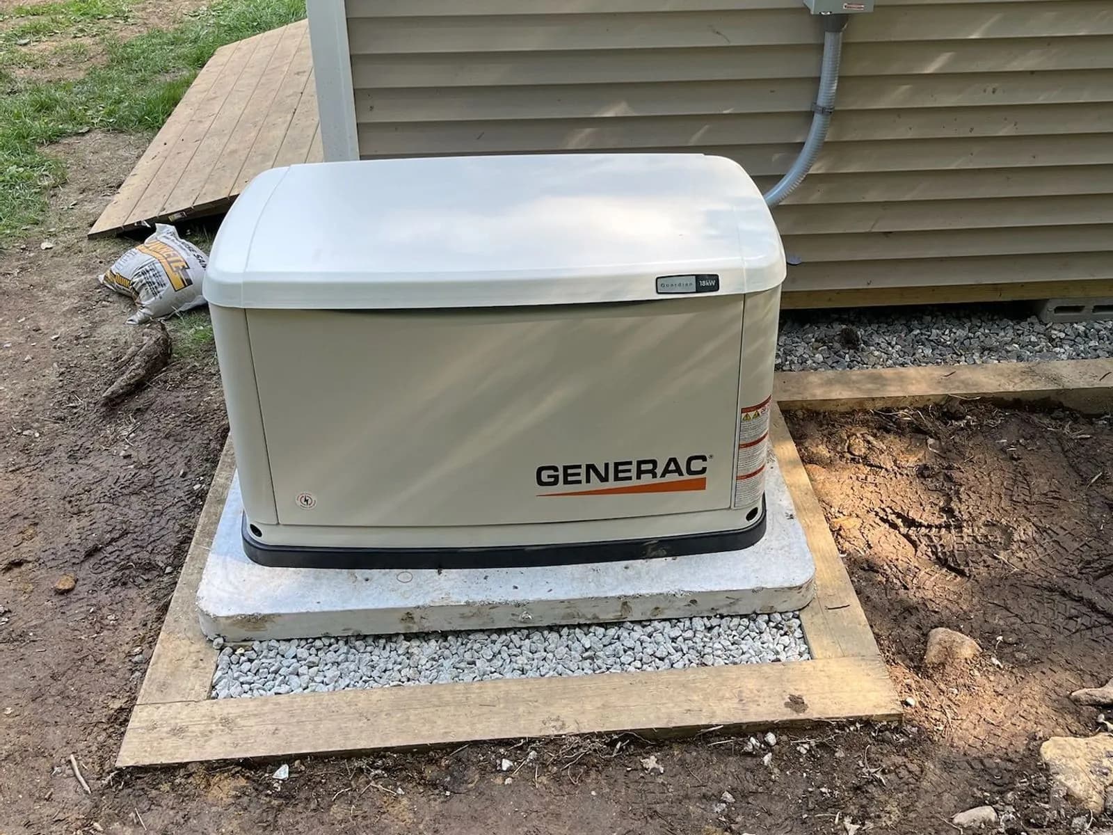 A new Generac standby generator on a concrete pad over a timber-framed gravel bed beside a tan vinyl-sided house, its conduit whip running up into a gray box on the wall.