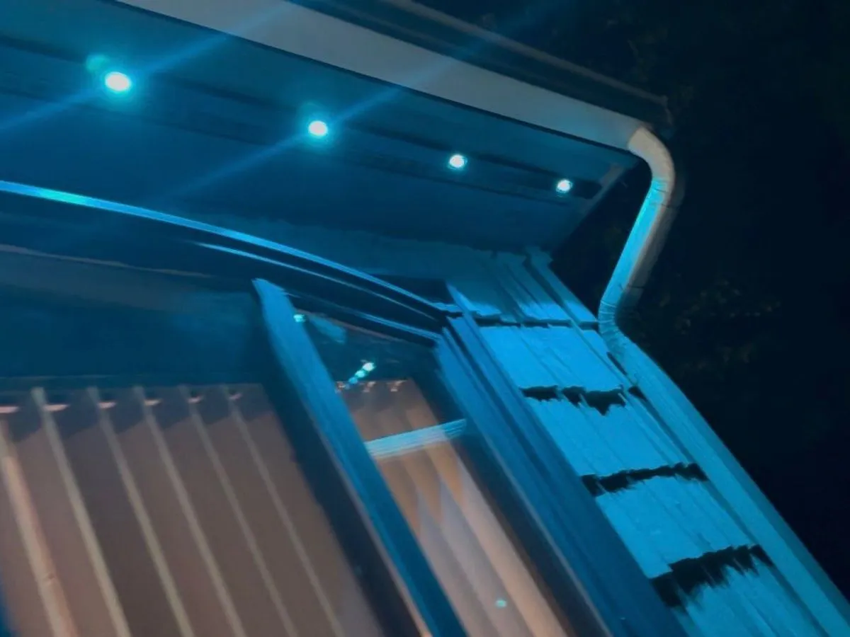 Teal permanent LED point lights set in a soffit glow at night, washing a curved bay window and shingled wall in blue-green light.