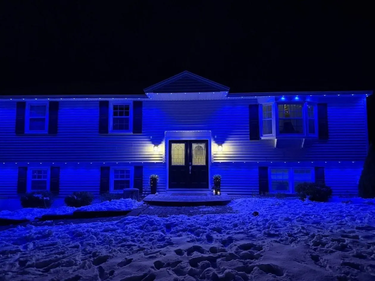 A raised ranch home at night bathed in deep blue from permanent eave lights, warm sconces beside the double front doors and footprints across the snowy lawn.