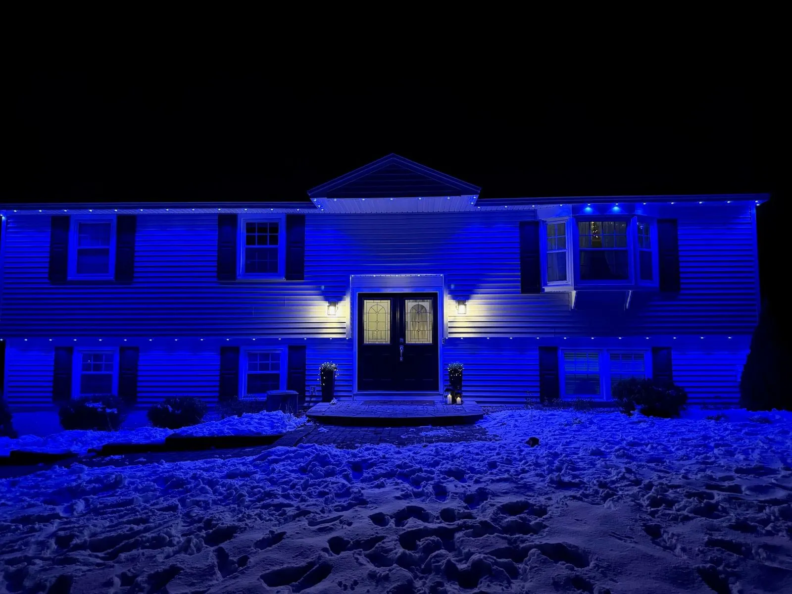 A split-level home at night in the snow, washed in deep blue from permanent eave lights, with warm sconces beside the double front doors.