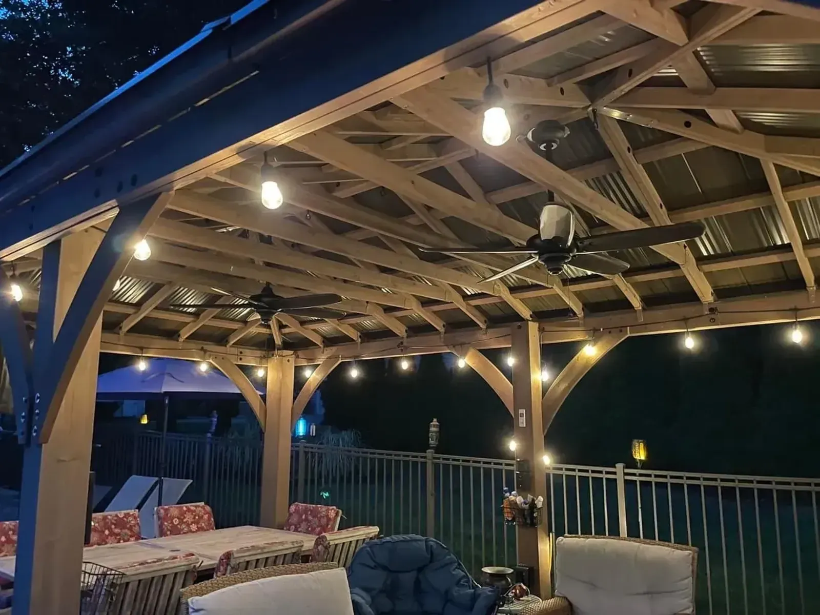 A timber pavilion at night strung with glowing bulb lights, ceiling fans in the rafters above outdoor seating, a pool fence beyond.
